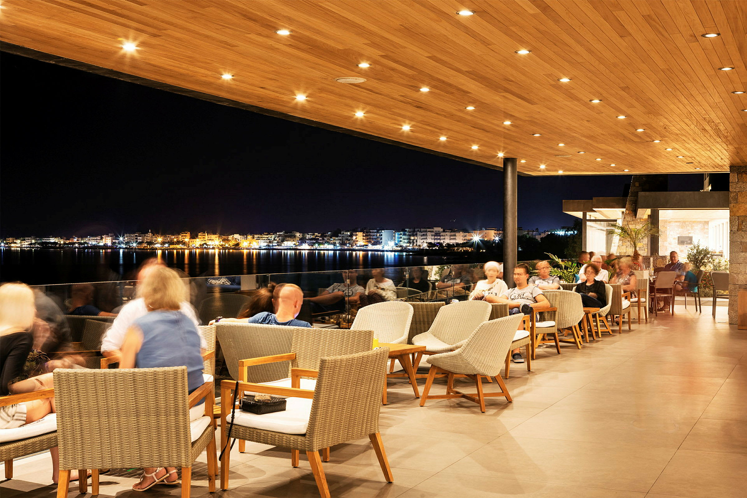 En restaurant med udendørsområde under en træpaneleret tagterrasse, med udsigt over en oplyst by ved vandet om aftenen. (AI-genereret tekst)