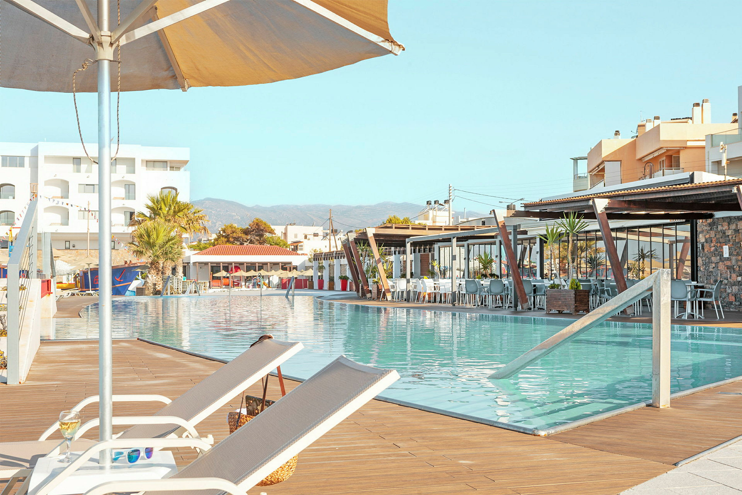 Billede af en solbeskinnet udendørs pool med liggestole og en parasol ved kanten, omgivet af bygninger og restauranter. (AI-genereret tekst)