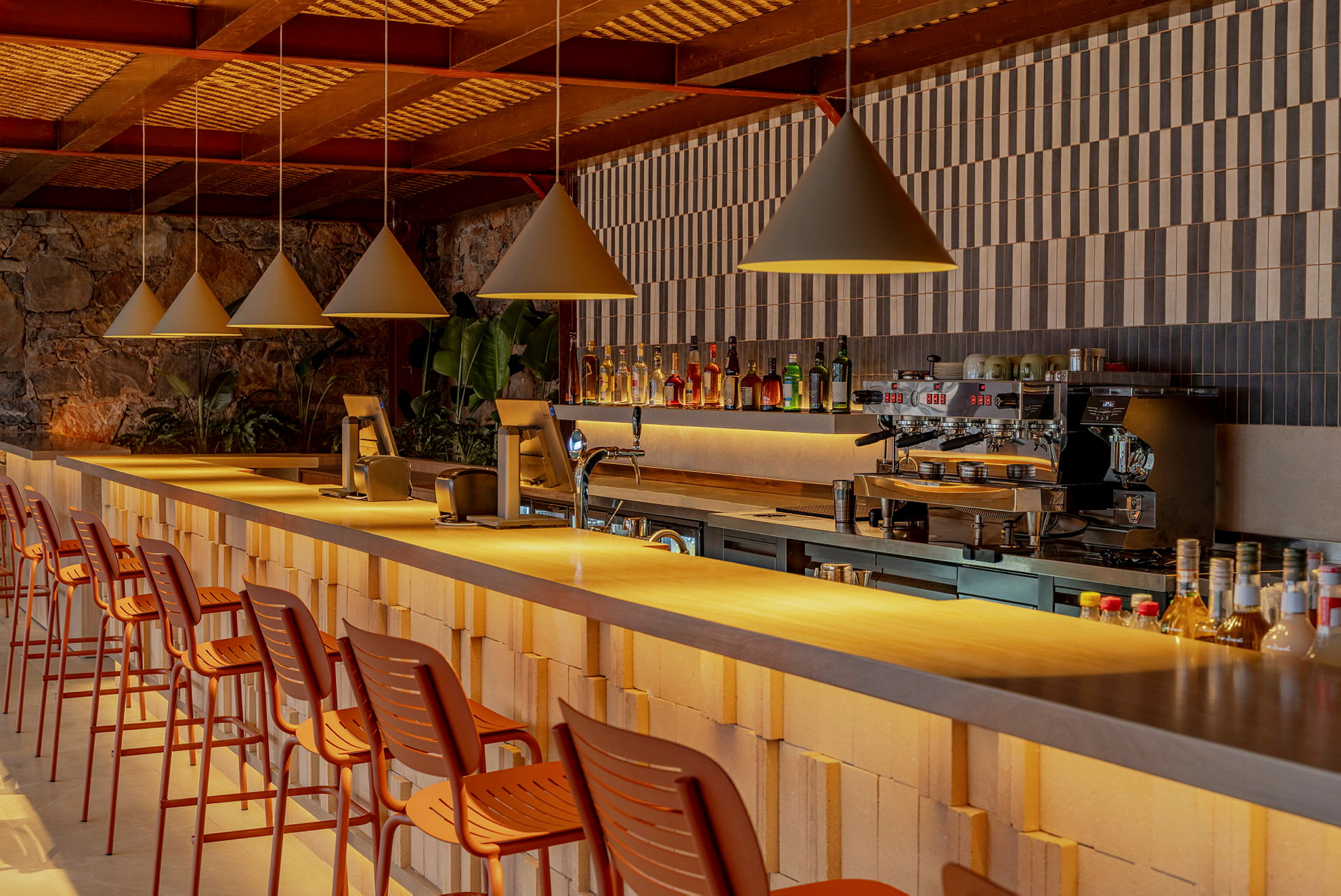 Billedet viser en moderne bar med en række høje stole ved en lang bardisk. Baren er dekoreret med flasker og har en kaffemaskine. (AI-genereret tekst)