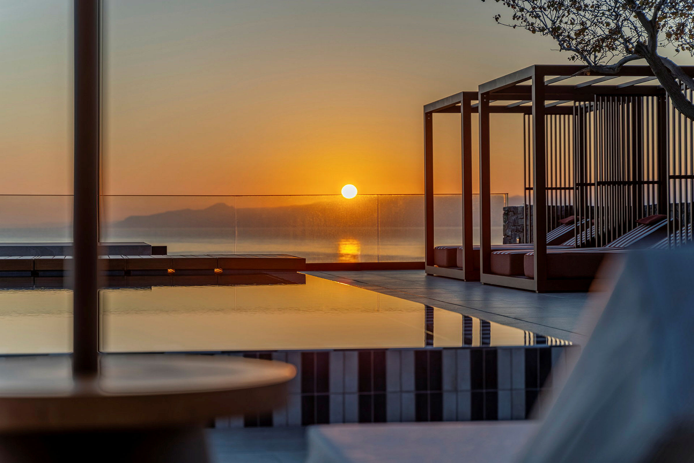 En solnedgang over en fjern horisont, reflekteres i en rolig pool omgivet af moderne møbler. (AI-genereret tekst)
