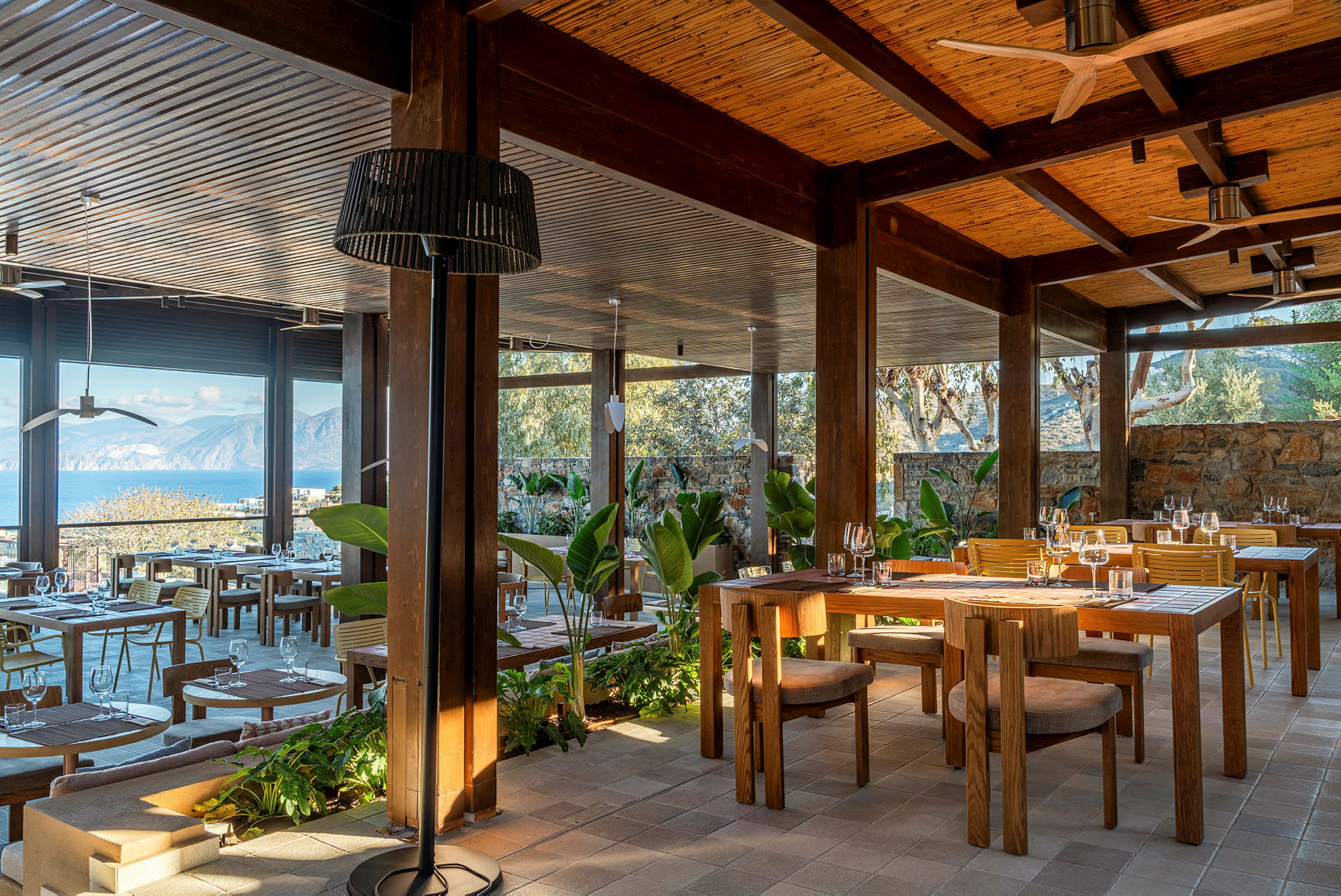 En udendørs restaurant med træmøbler og udsigt over havet og bjergene. (AI-genereret tekst)