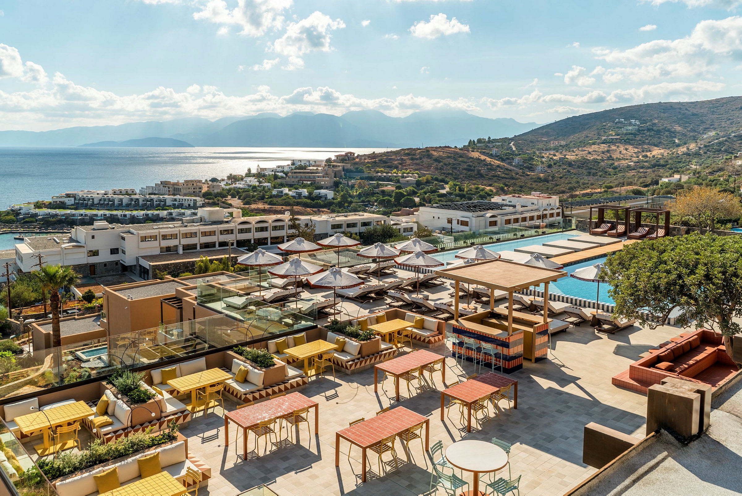 Billede af et hotelresort med en solrig terrasse, poolområde og udsigt over havet og bakker. (AI-genereret tekst)