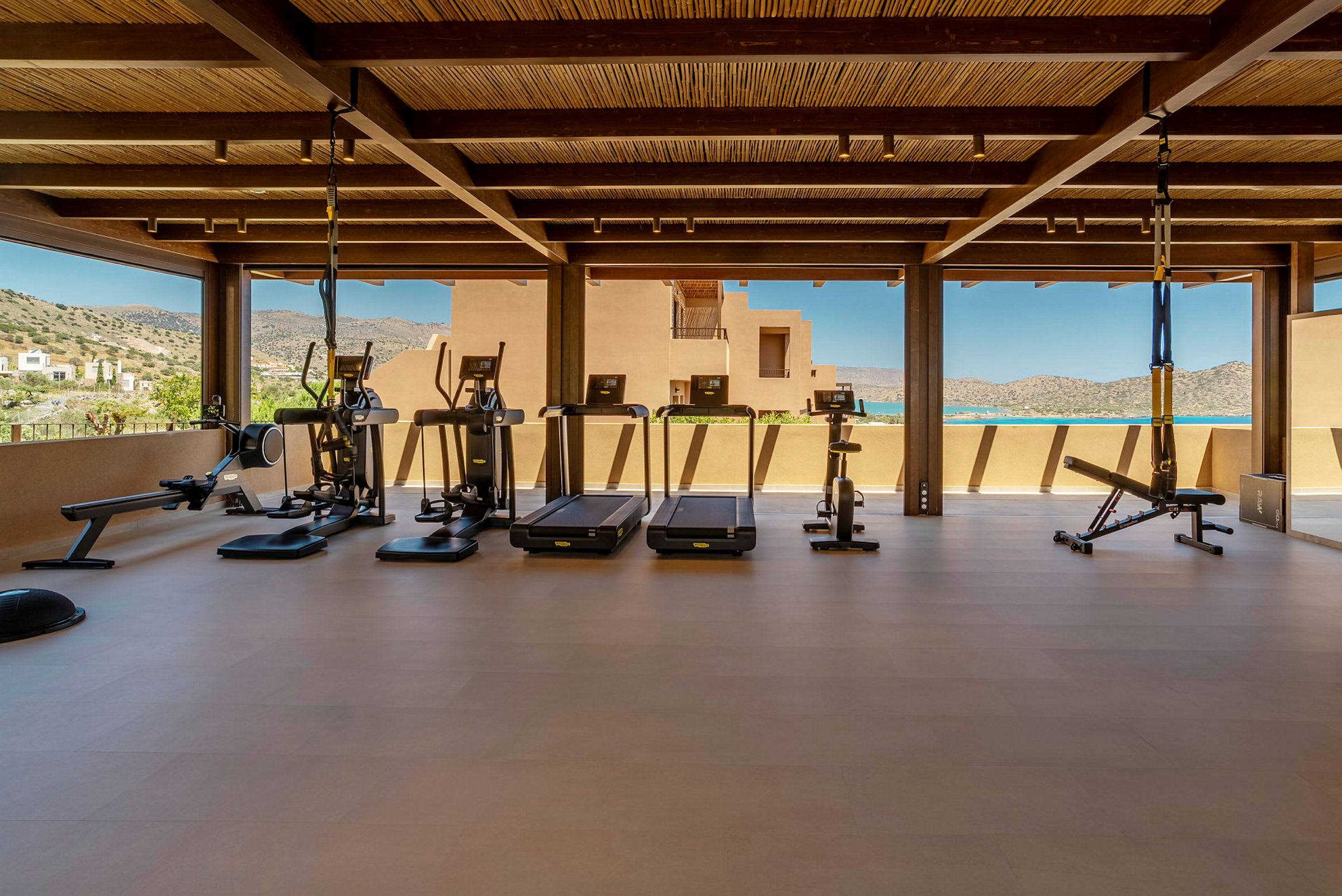 Et fitnesscenter med træningsudstyr, såsom løbebånd, motionscykler og styrketræningsredskaber, under et tag med udsigt over bjergene. (AI-genereret tekst)