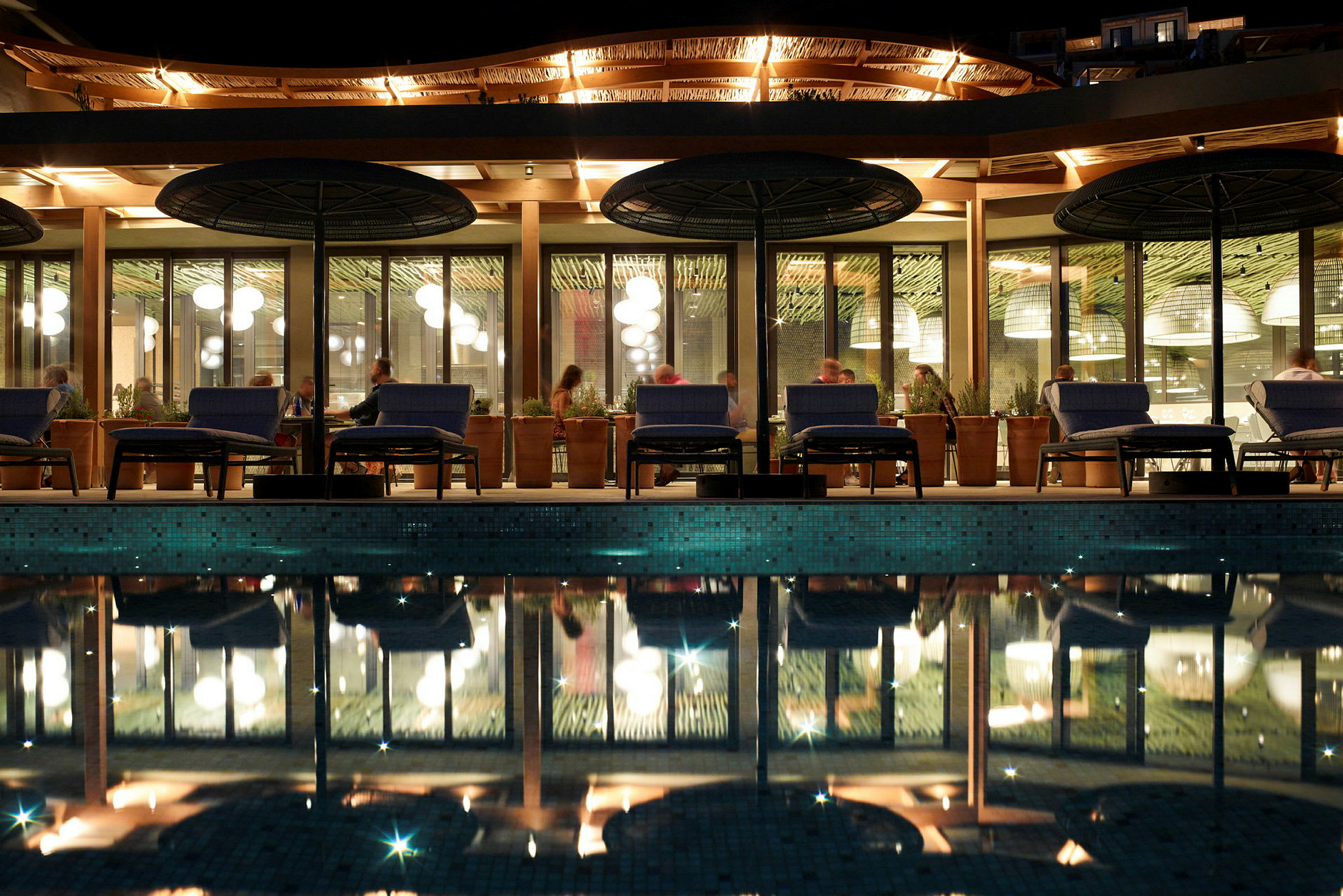 Billede af udendørsområdet ved en restaurant med aftenbelysning, en pool i forgrunden og gæster ved borde. (AI-genereret tekst)
