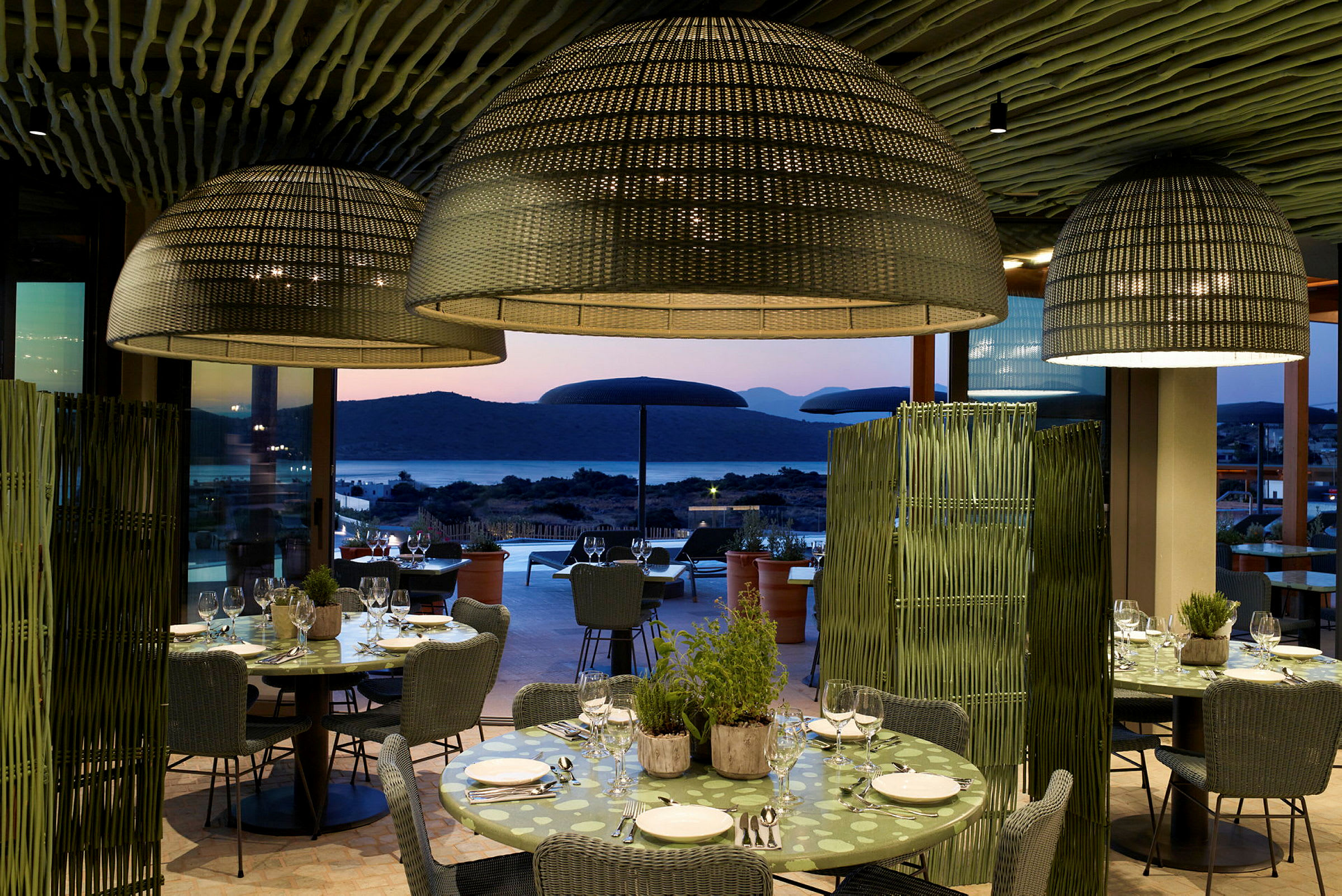 Restaurant med dækkede borde og store lampeskærme, udsigt mod havet i solnedgangen. (AI-genereret tekst)