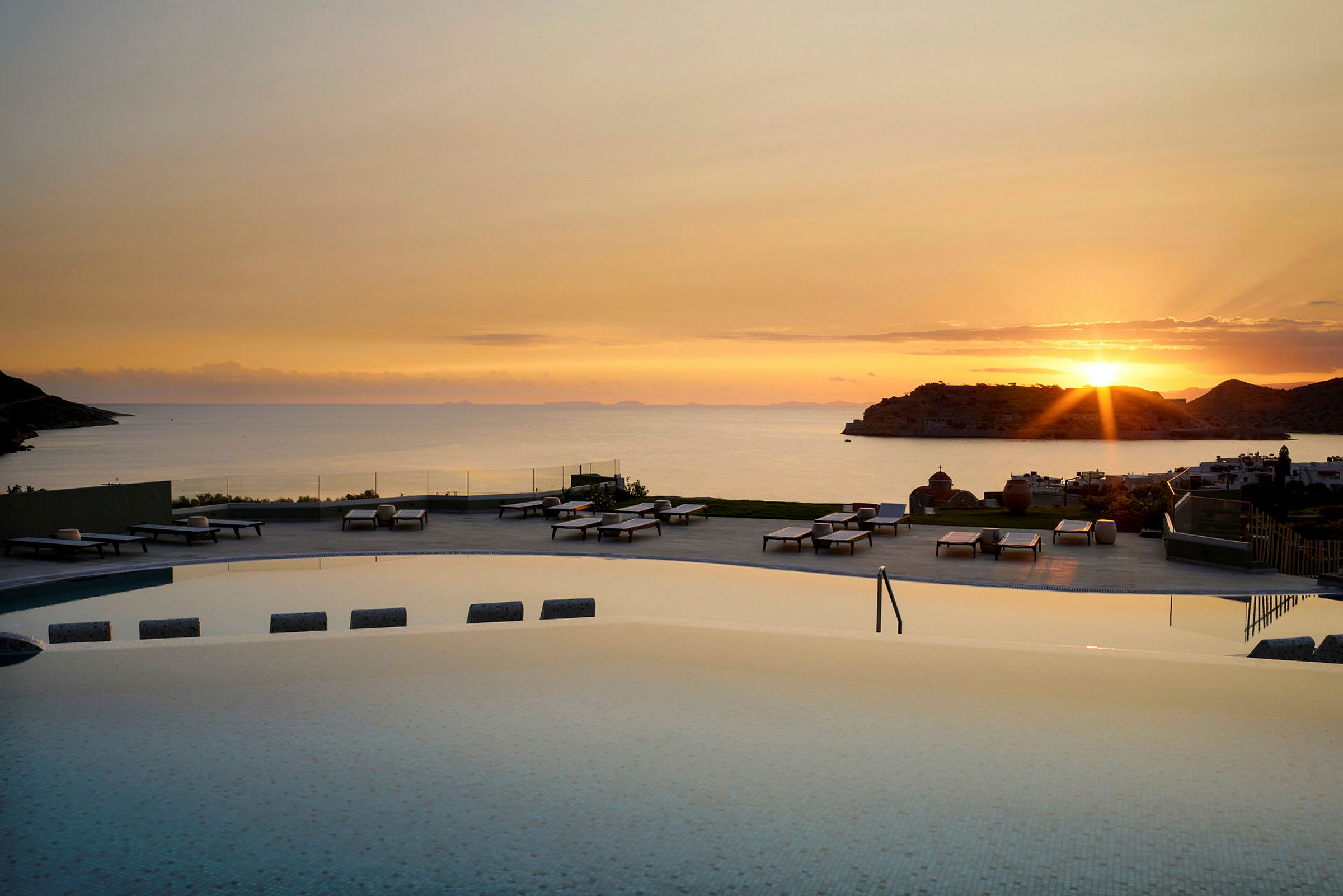 Udsigt over en pool med liggestole i forgrunden og havet ved solnedgang i baggrunden. (AI-genereret tekst)