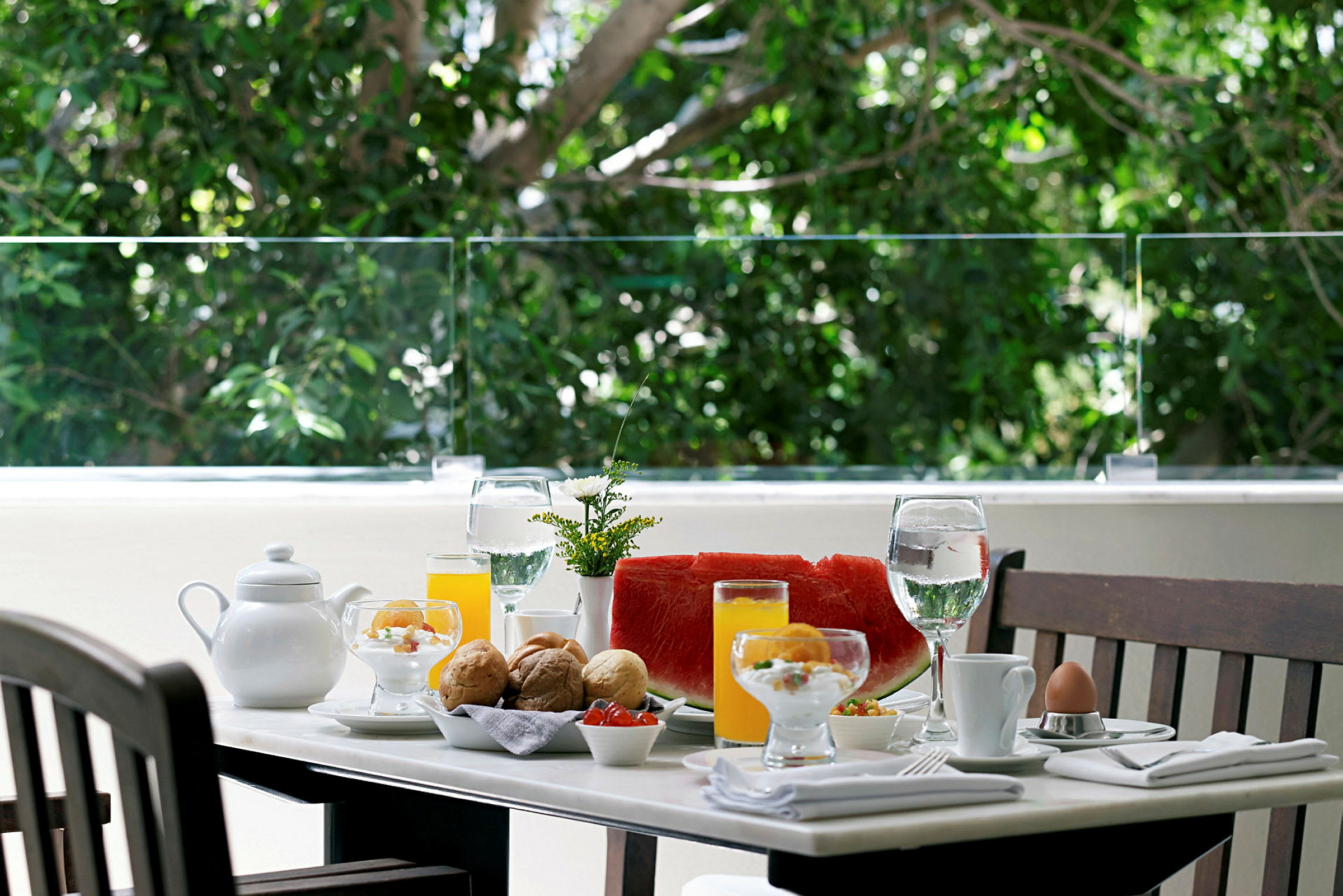 Et udendørs bord med morgenmad serveret, herunder glas, juice, frugt og en tekande. (AI-genereret tekst)