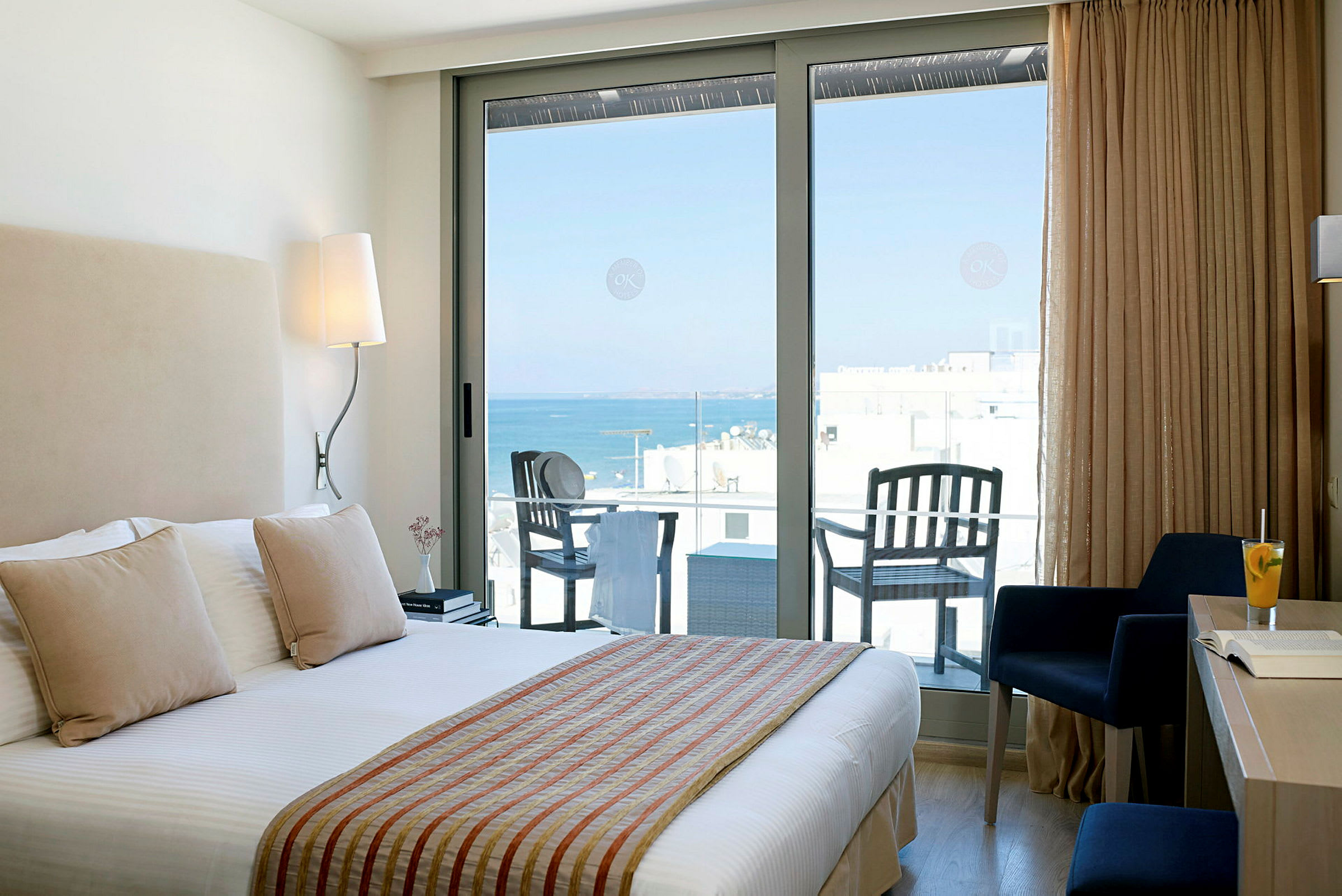 Hotelværelse med seng og vindue ud til en altan med udsigt over havet. (AI-genereret tekst)