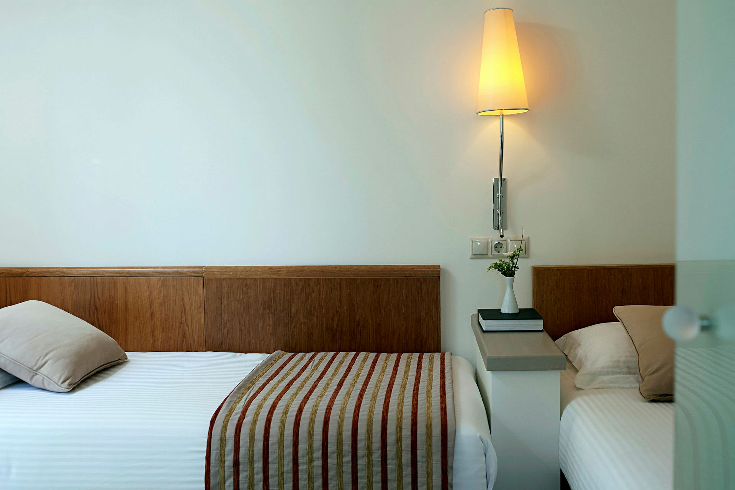To enkeltsenge på et hotelværelse med træhovedgærder og hvidt sengetøj med stribede sengetæpper. En lampe og en lille vase med blomster står på et bord mellem sengene. (AI-genereret tekst)