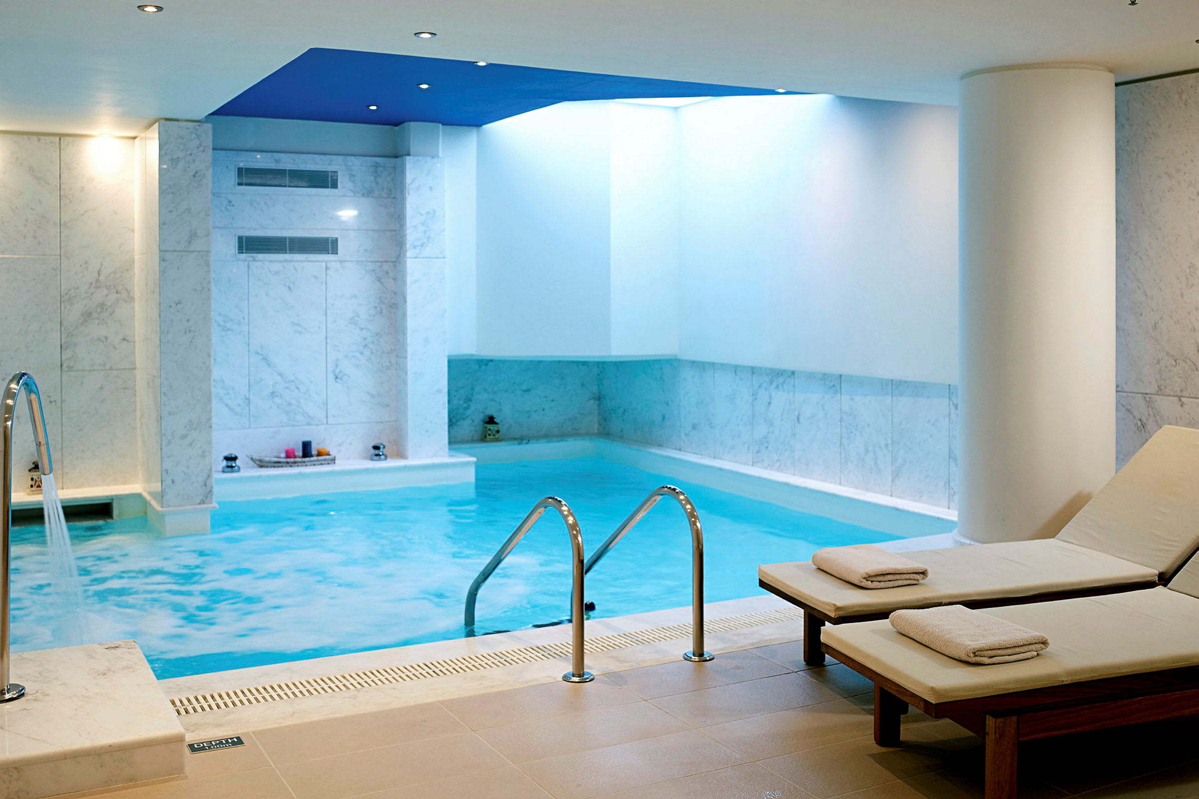 Et indendørs poolområde med pool, to liggestole med håndklæder og hvide flisevægge. (AI-genereret tekst)