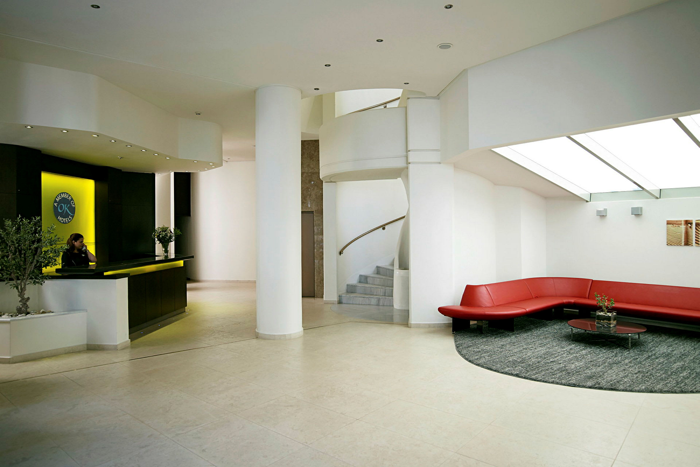 Et moderne hotellreceptionsområde med en receptionsskranke, et cirkelformet siddeområde med røde sofaer og en trappe. (AI-genereret tekst)