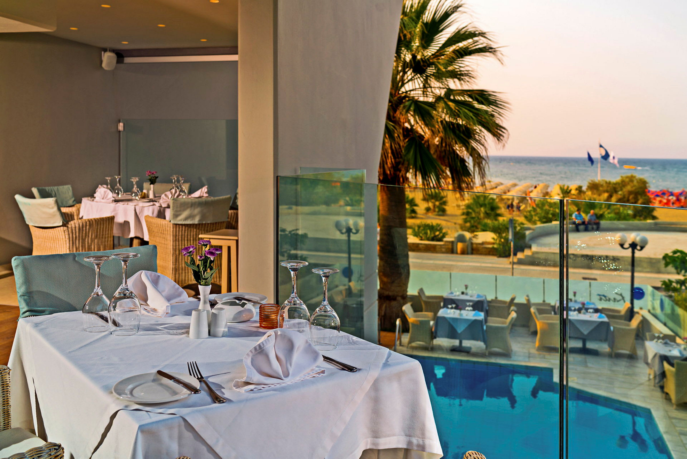 Et elegant dækket bord på en restaurantterrasse med udsigt over en pool, palmer og stranden i baggrunden. (AI-genereret tekst)