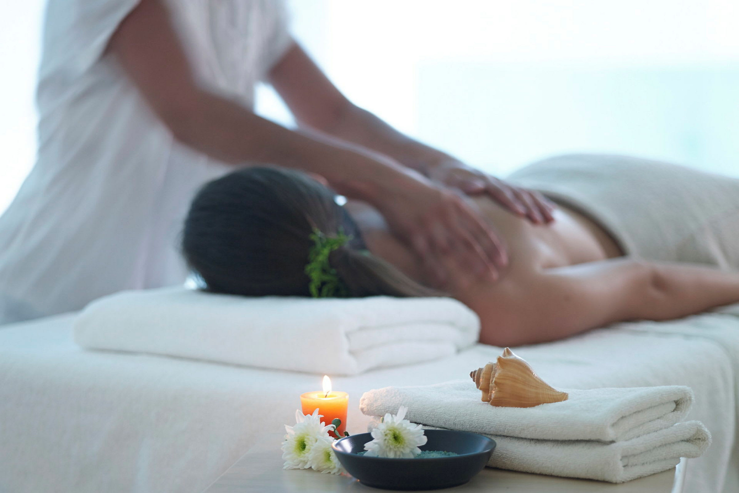 En person får en rygmassage på et spa. I forgrunden ses en flamme, blomster og en konkylie-dekoration. (AI-genereret tekst)