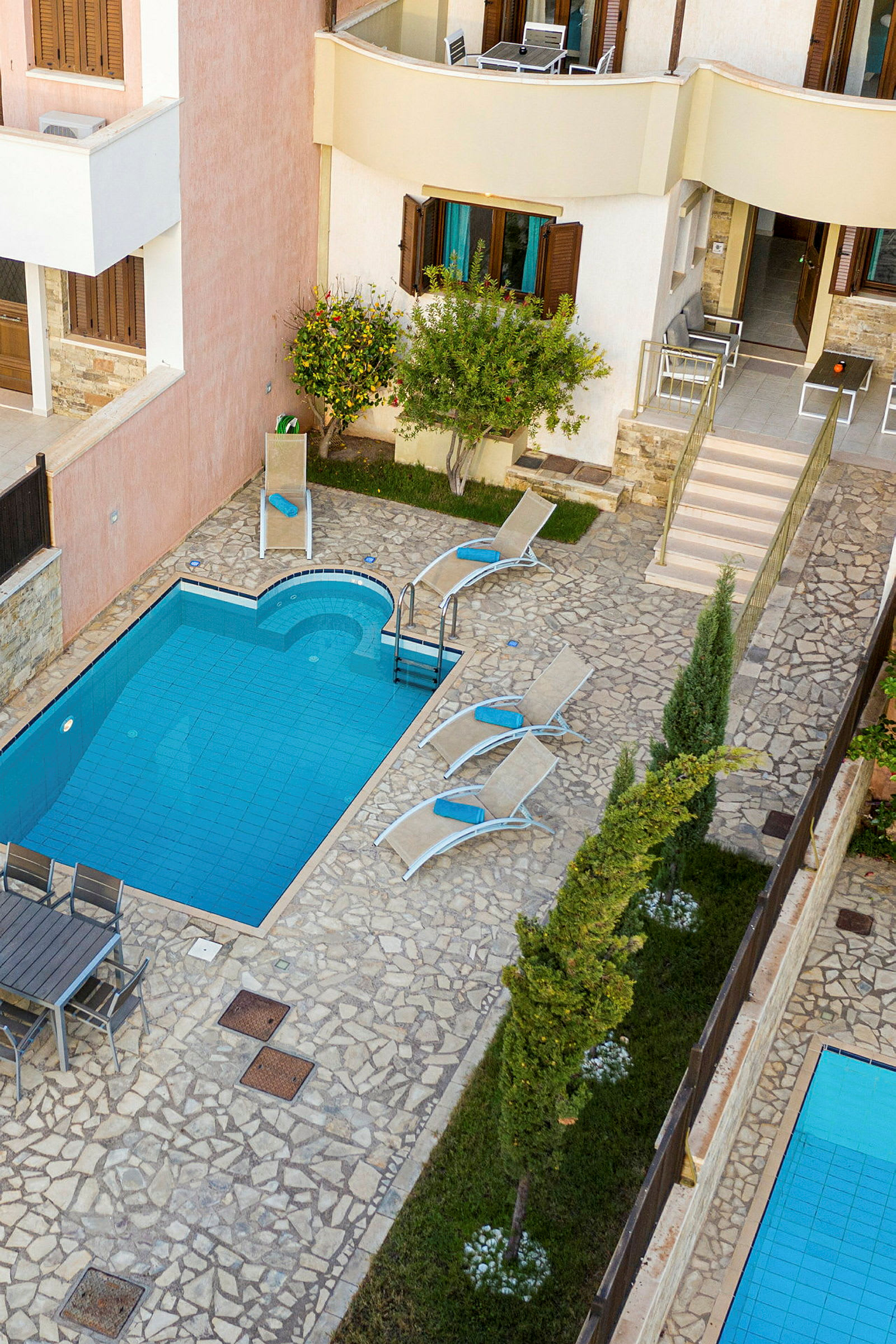 En lille pool ved en brostensbelagt terrasse med liggestole og grønne træer omkring. (AI-genereret tekst)