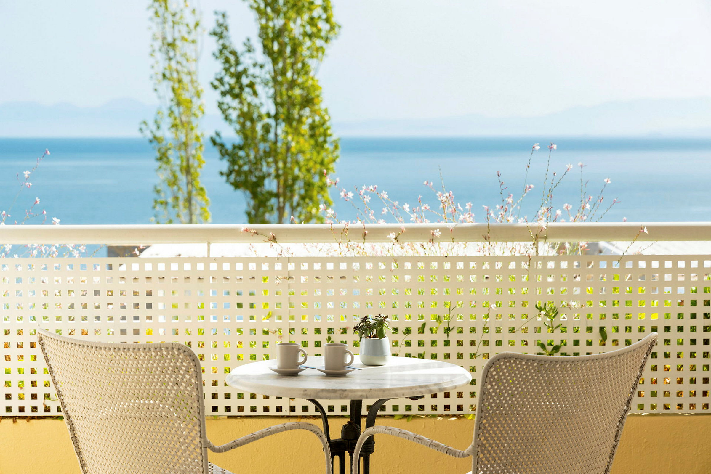 Udsigt fra en terrasse med et bord og to stole, blomster og hav i baggrunden. (AI-genereret tekst)