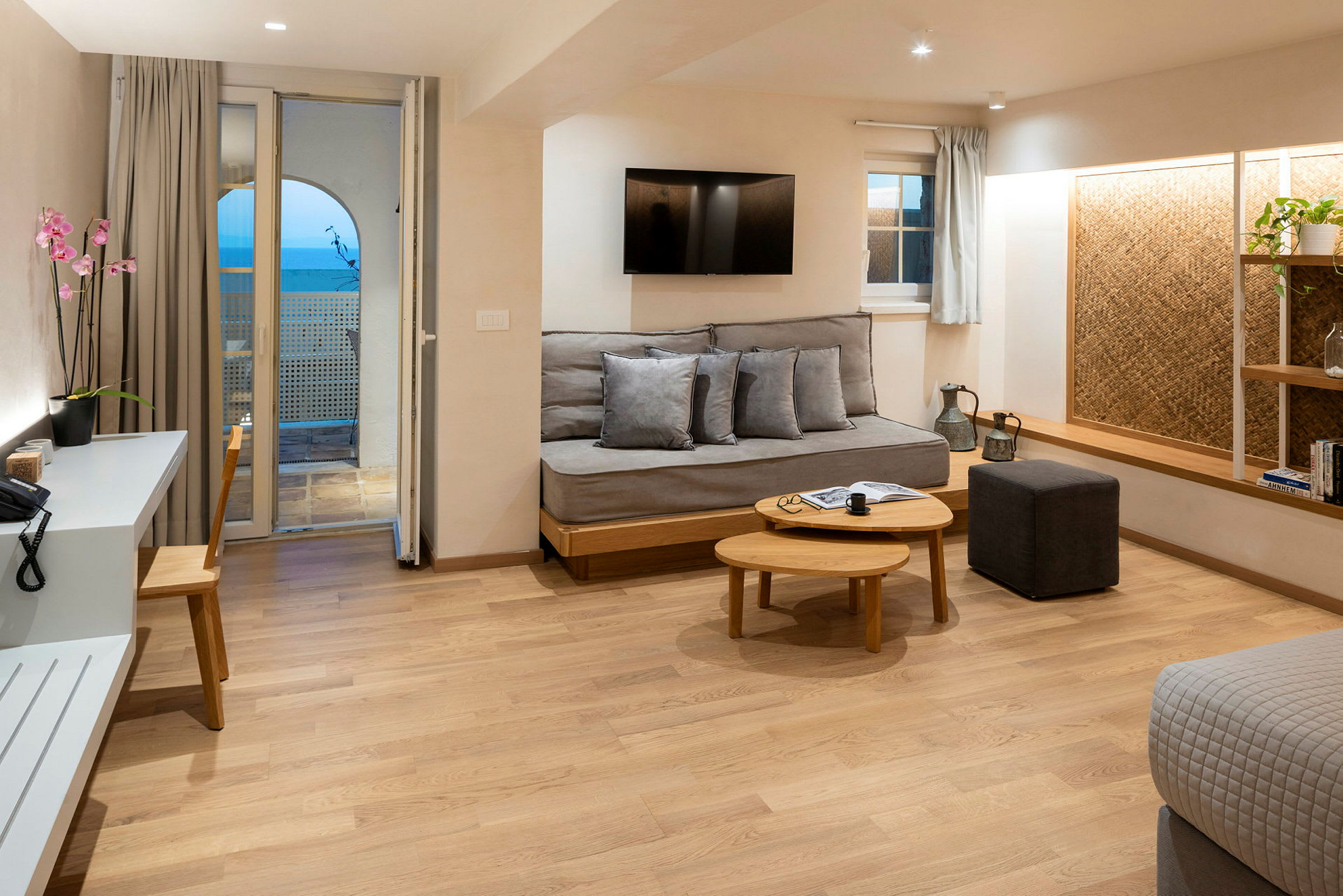 Et moderne hotelværelse med trægulv, sofa, sofabord, skrivebord med stol samt en altan med havudsigt. (AI-genereret tekst)