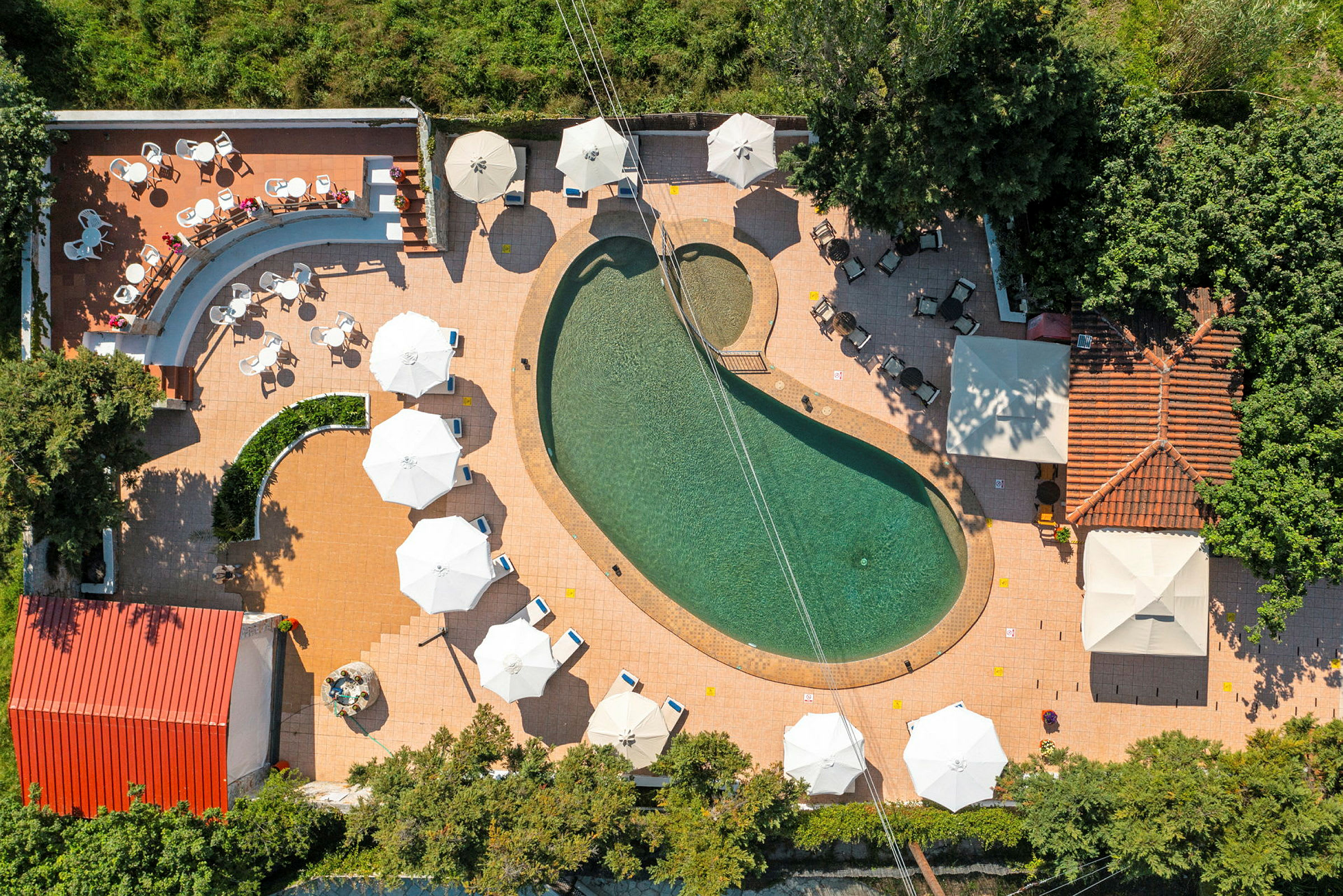 Luftfoto af en oval swimmingpool omgivet af parasoller og siddepladser. (AI-genereret tekst)