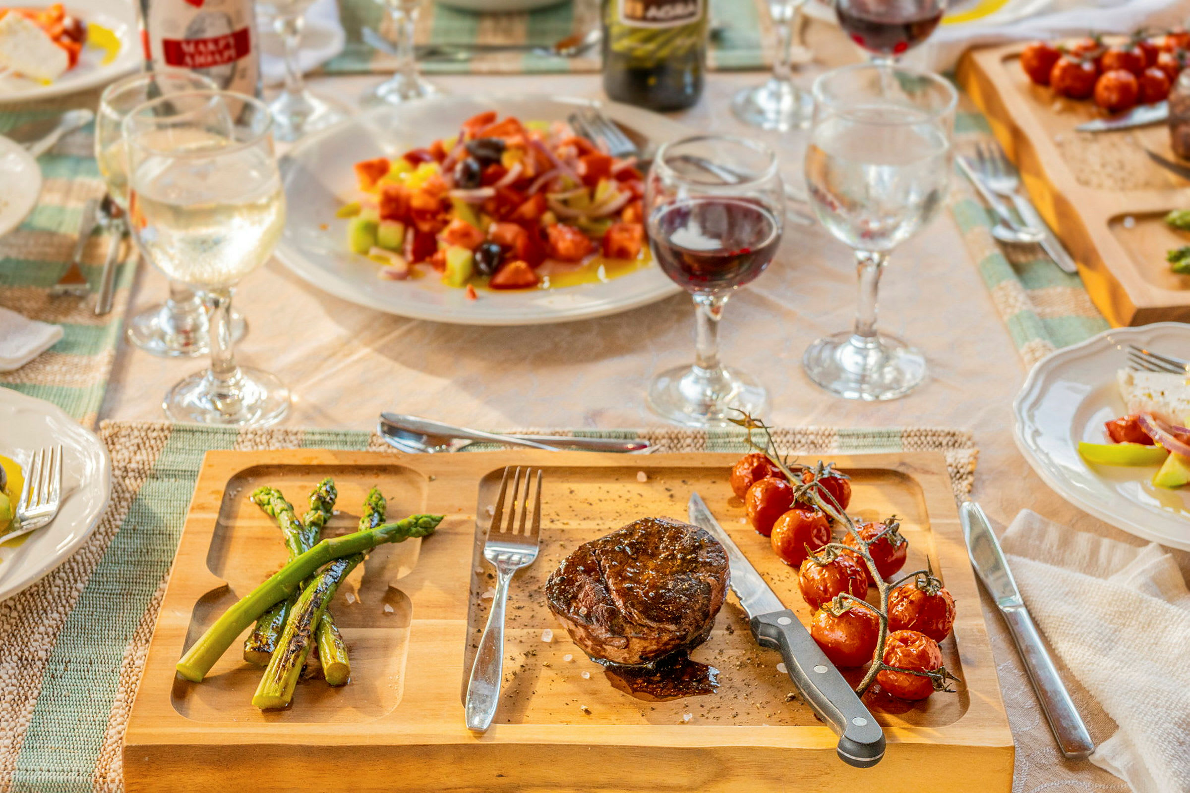 En træbakke med en portion grillet kød, asparges og cocktailtomater, omgivet af dækket bord med glas og tallerkener. (AI-genereret tekst)