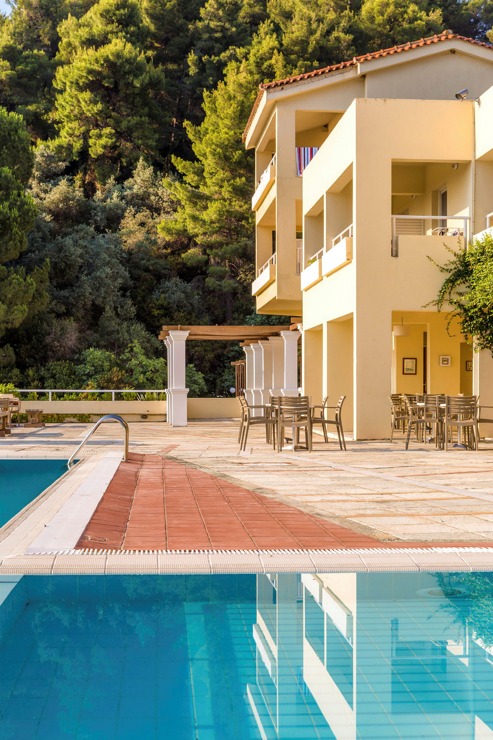 Hotellets poolområde med borde og stole samt skov i baggrunden. (AI-genereret tekst)
