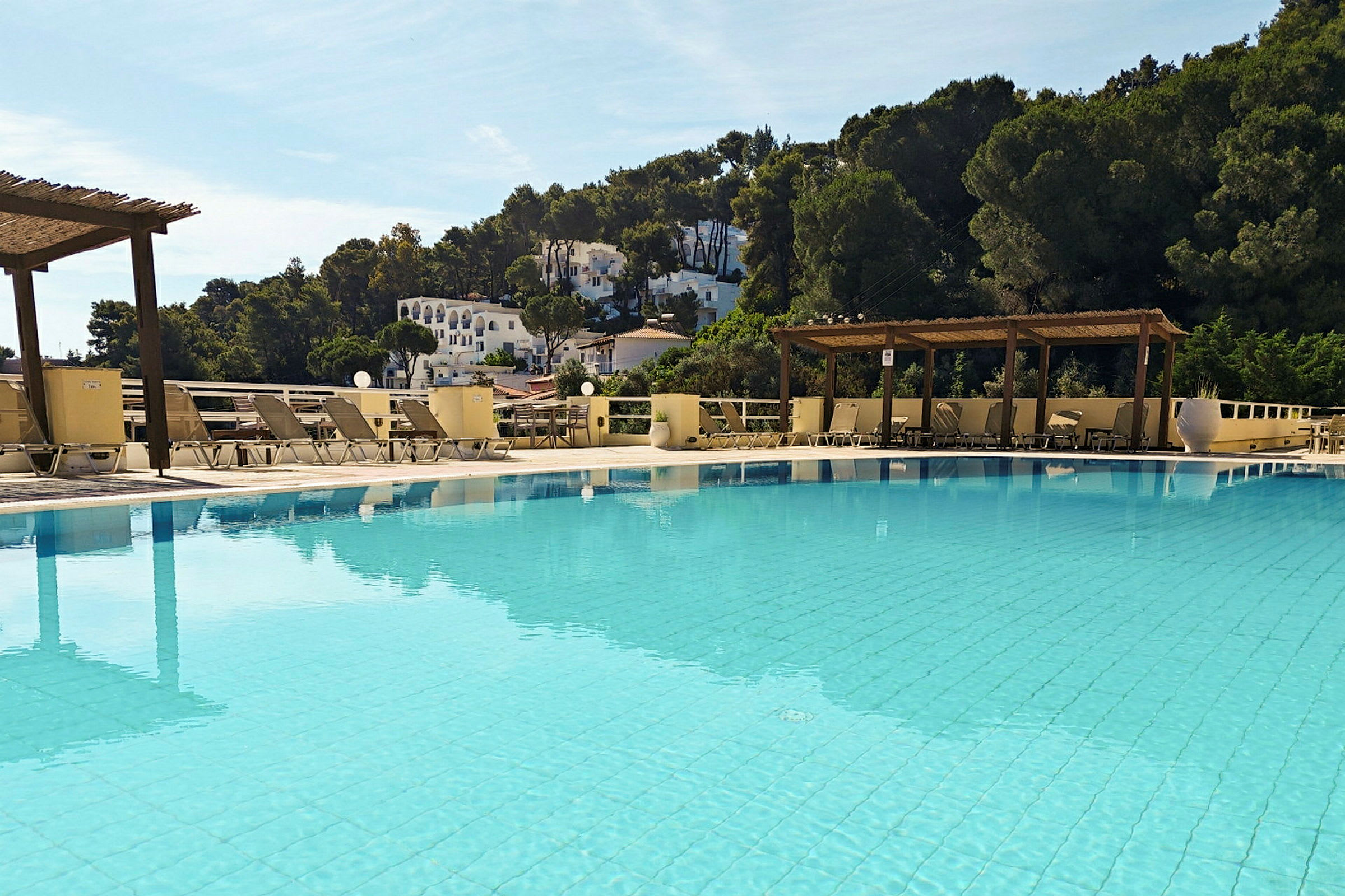 En udendørs swimmingpool omgivet af liggestole og pergola. I baggrunden er der træer og hvide bygninger på en skråning. (AI-genereret tekst)