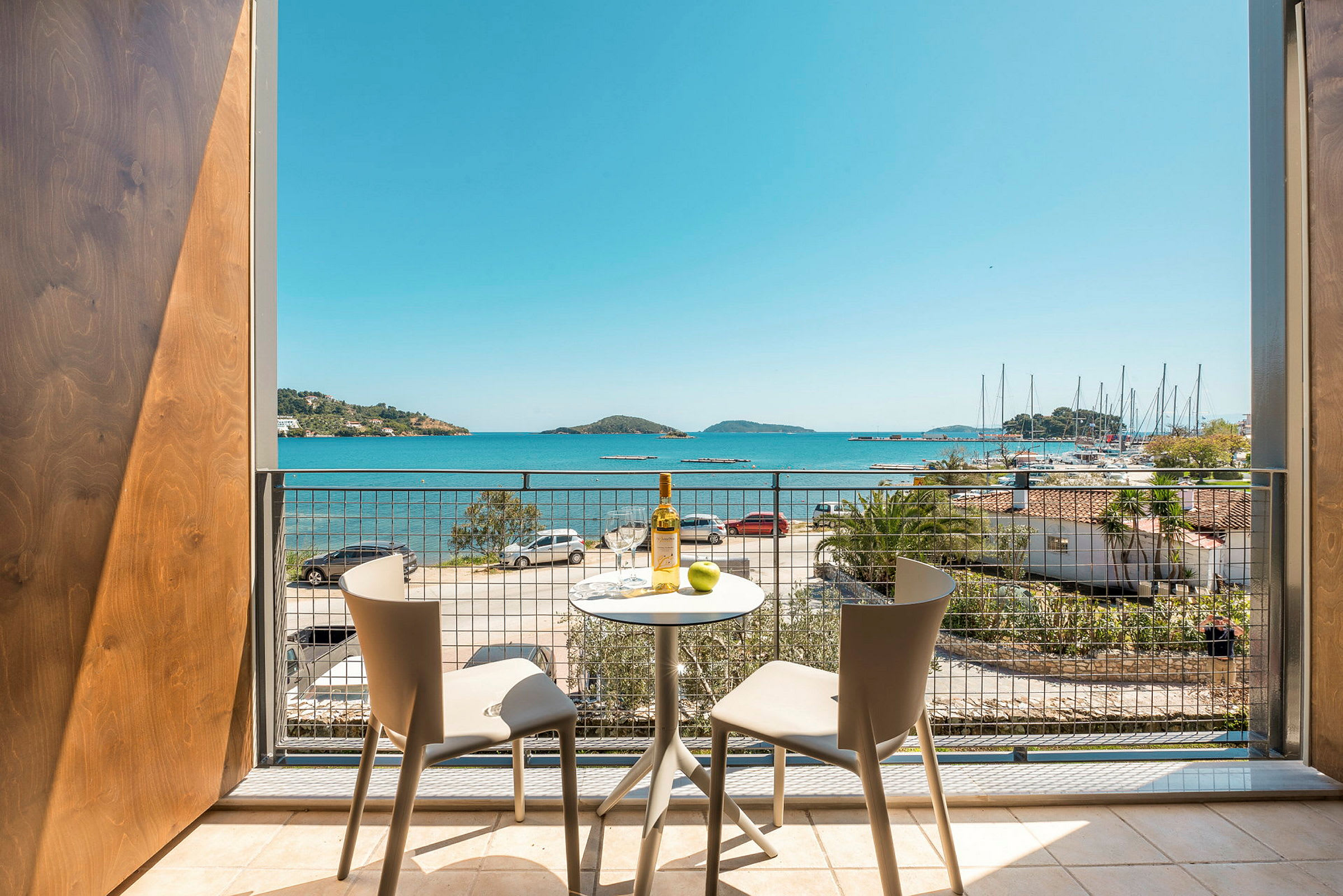 Udsigt fra en balkon med to stole og et bord, mod havet og en lystbådehavn. (AI-genereret tekst)
