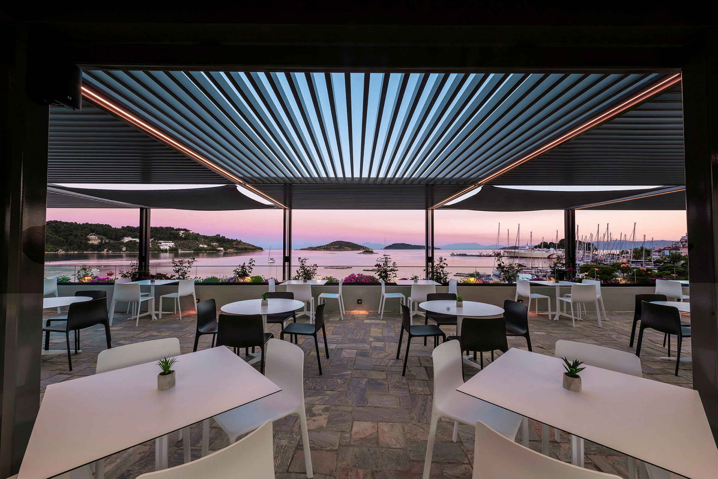 En udendørs terrasse med borde og stole med udsigt over havet og sejlbåde. (AI-genereret tekst)