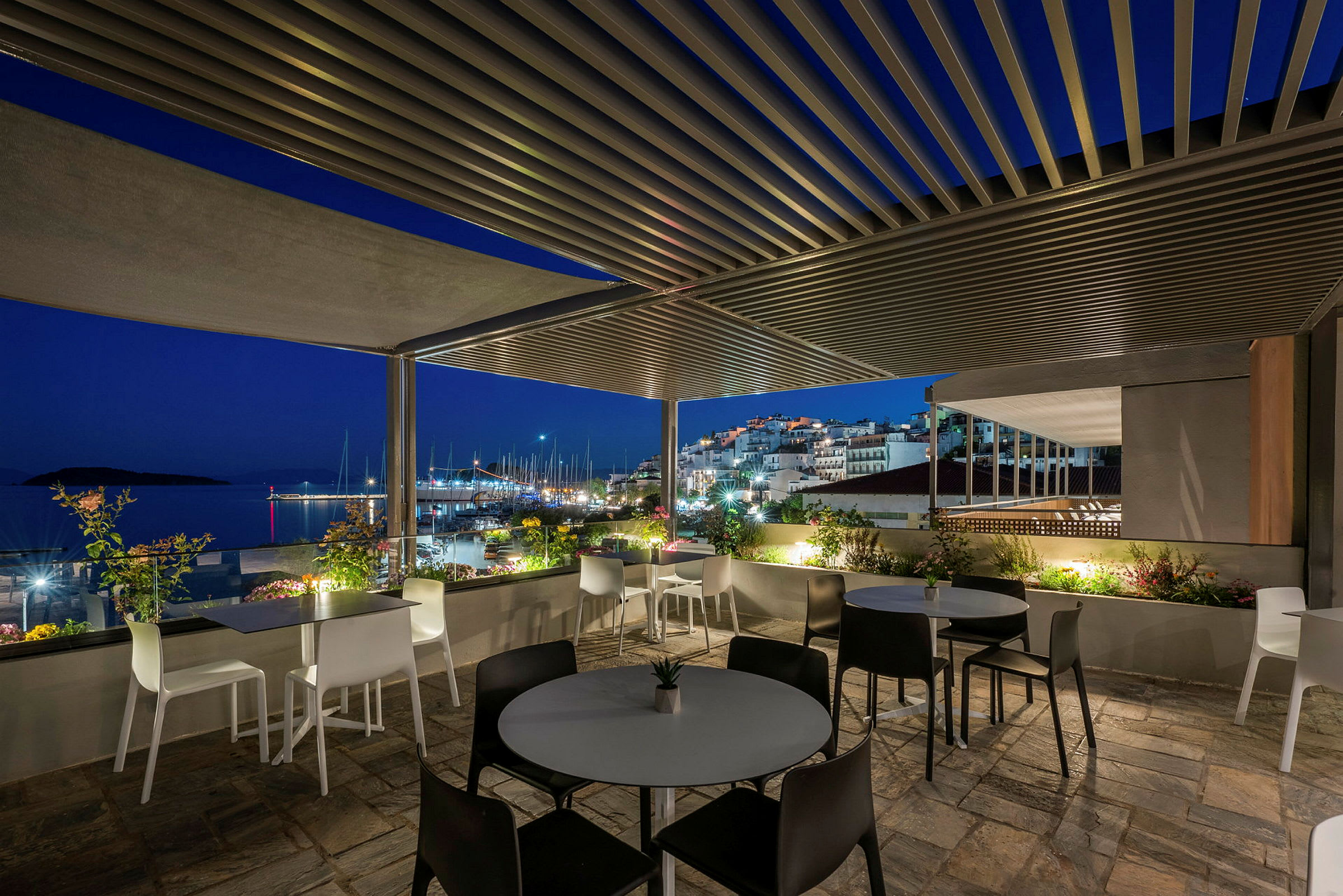 En terrasse med siddepladser og udsigt over en lystbådehavn og bygninger om aftenen. (AI-genereret tekst)