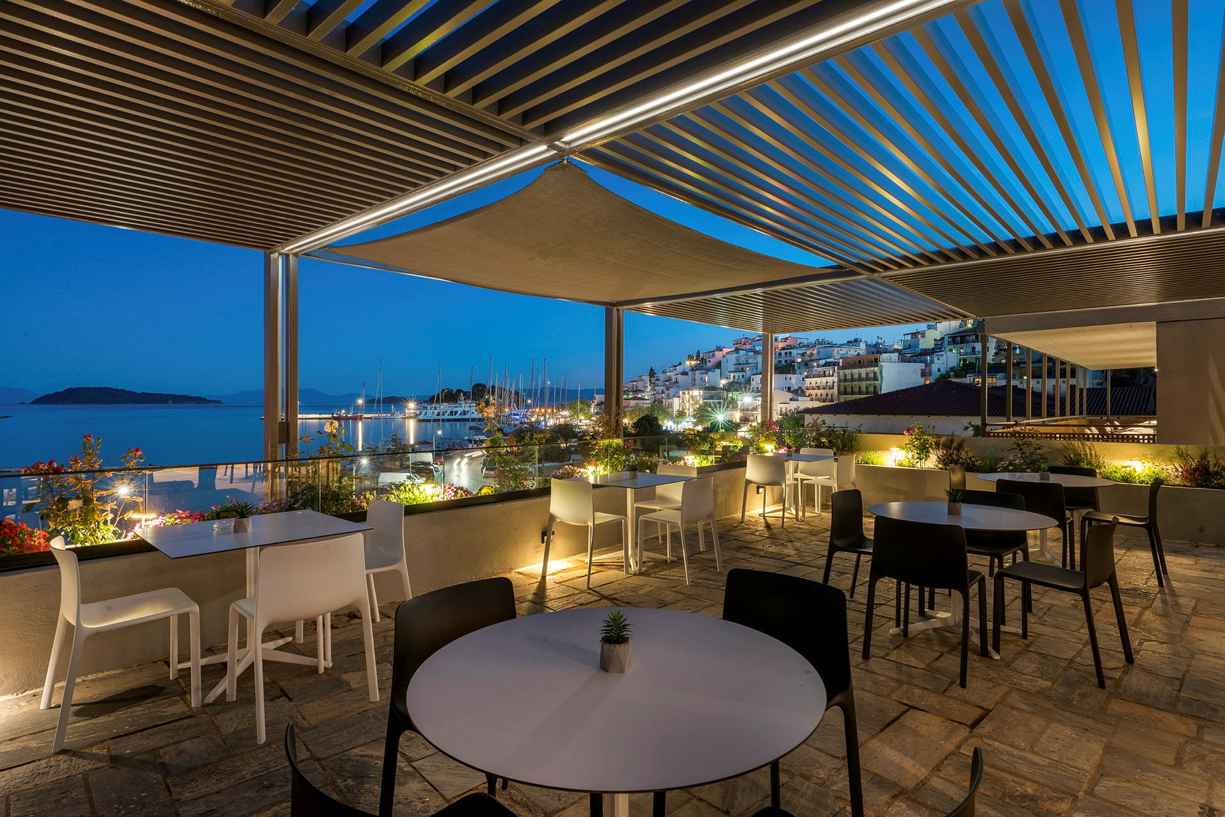 En udendørs terrasse med borde og stole under en pergola, med udsigt til en småbådshavn og en oplyst by ved kysten i tusmørket. (AI-genereret tekst)