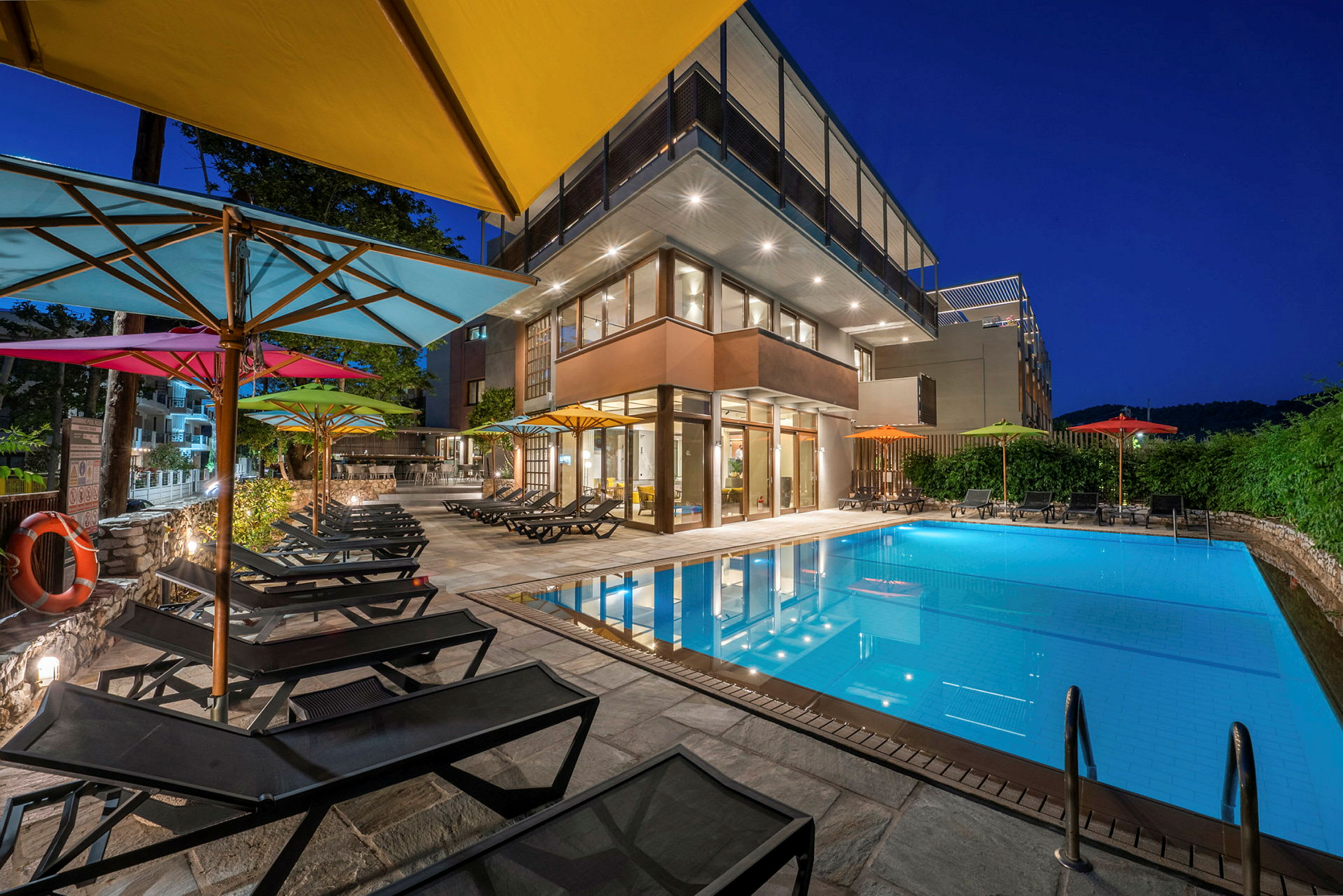 En udendørs pool med liggestole og farverige parasoller ved et moderne hotel om aftenen. (AI-genereret tekst)
