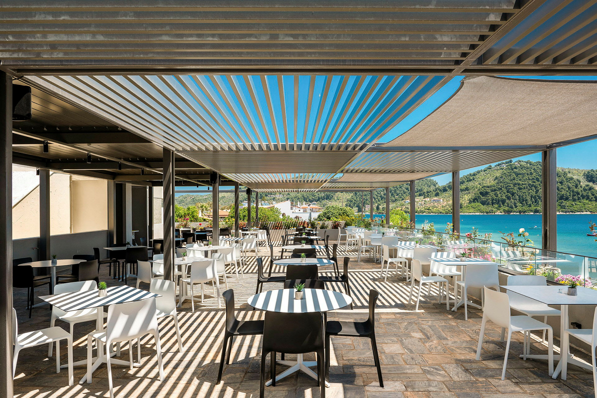 En udendørsrestaurant med hvide og sorte stole og borde under solafskærmning, med udsigt over havet og omgivelser med grønt. (AI-genereret tekst)