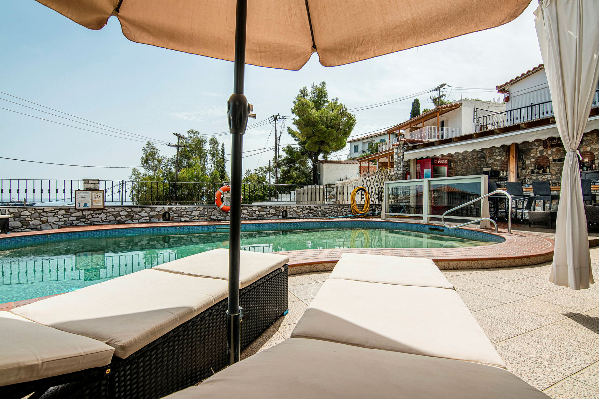 Billede af en pool med omkringliggende terrasse og liggestole under en parasol, samt en bygning og grønne områder i baggrunden. (AI-genereret tekst)