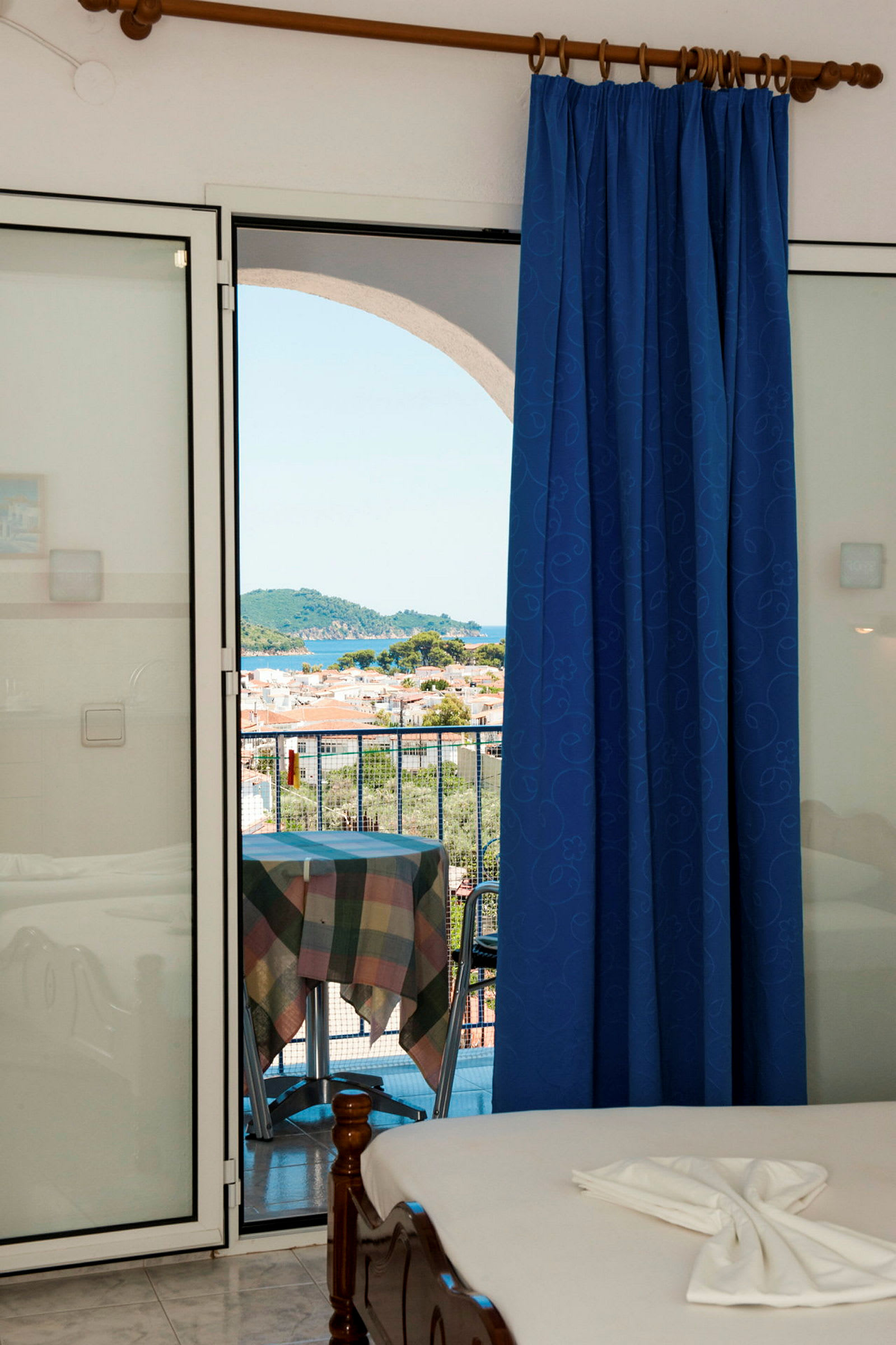 Udsigt fra et hotelværelse med en blå gardin og udgang til en balkon, der viser en malerisk by og natur. (AI-genereret tekst)