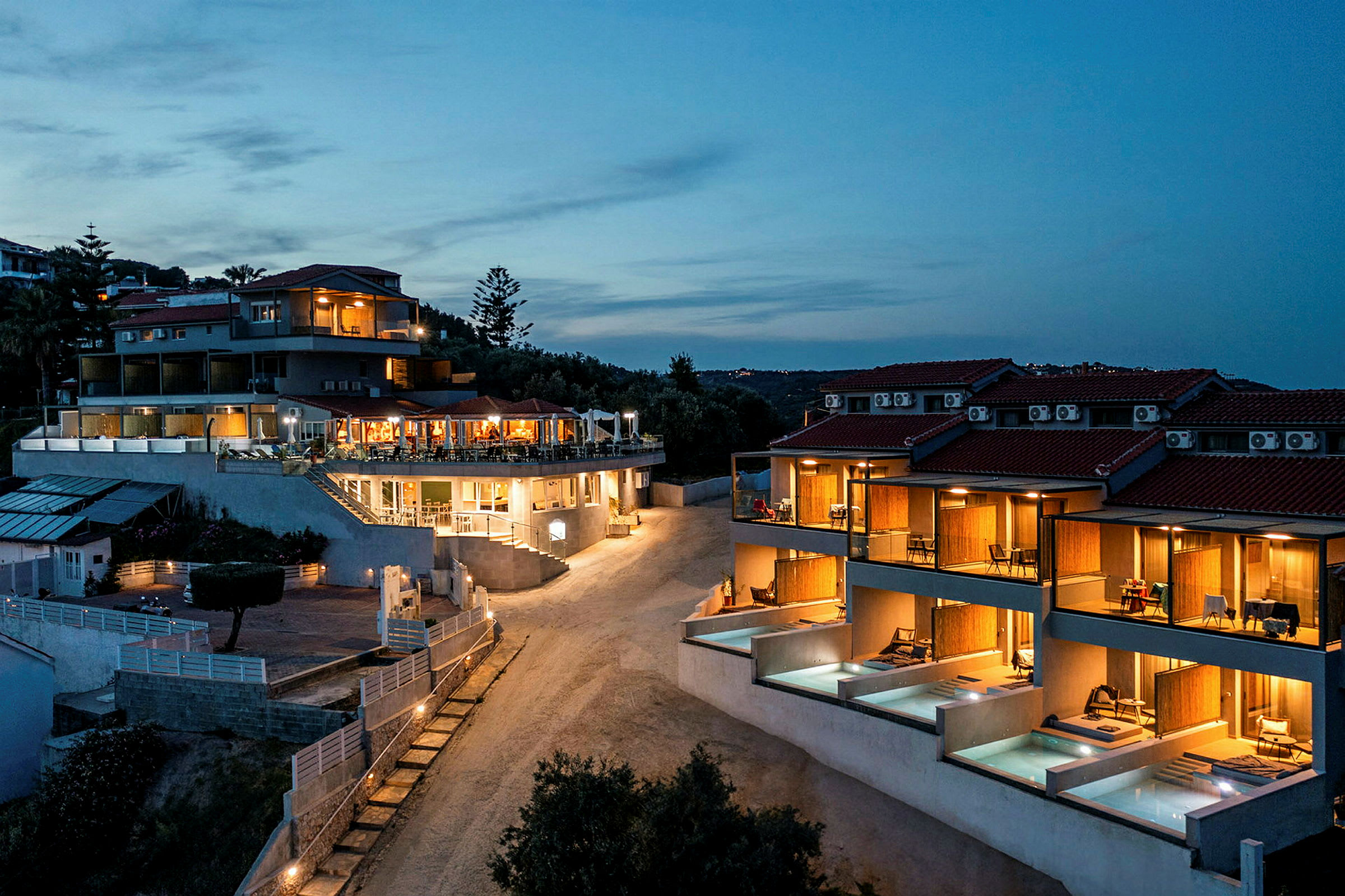 Billede af et hotel med oplyste bygninger og private poolområder under en aftenhimmel. (AI-genereret tekst)