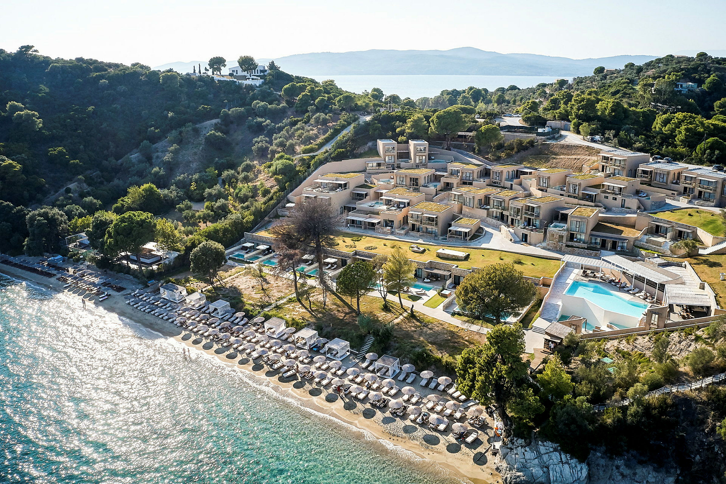 Luftfoto af resort med pool, strand med parasoller og liggestole samt omkringliggende beplantning. (AI-genereret tekst)