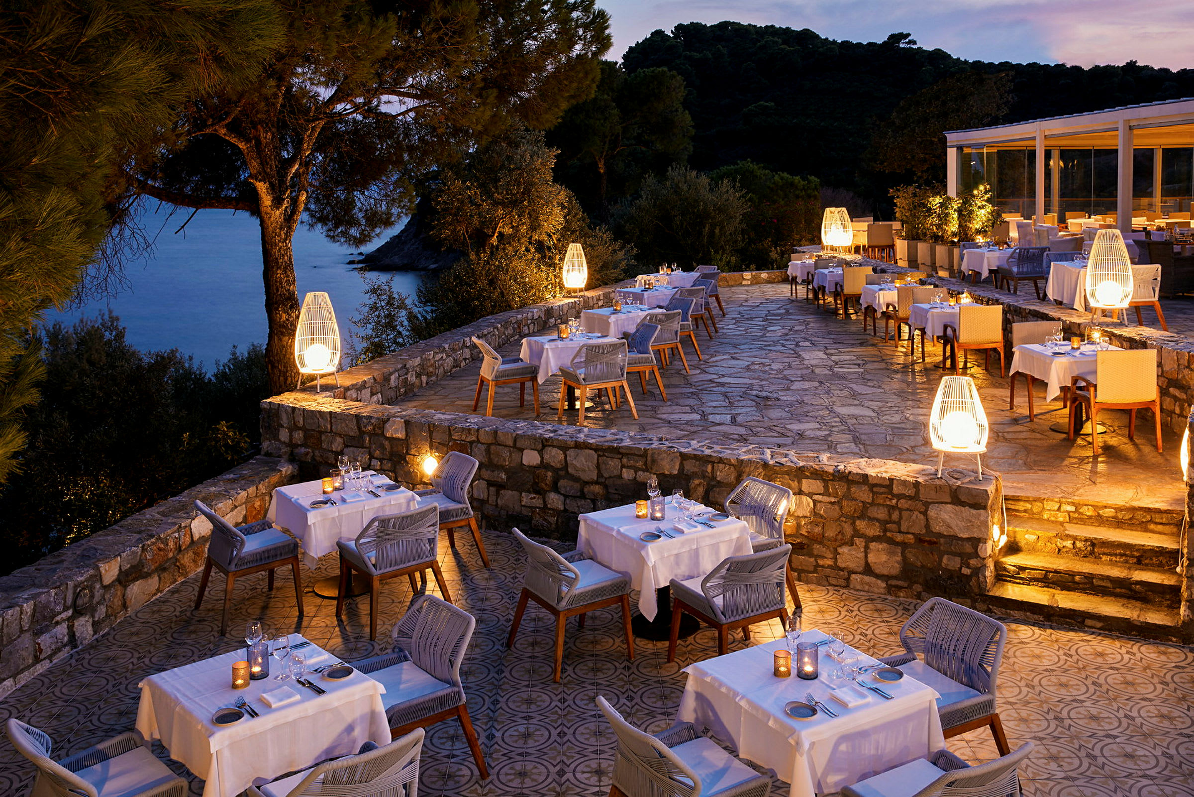 En restaurantterrasse med belysning, dækkede borde og udsigt over havet ved solnedgang. (AI-genereret tekst)