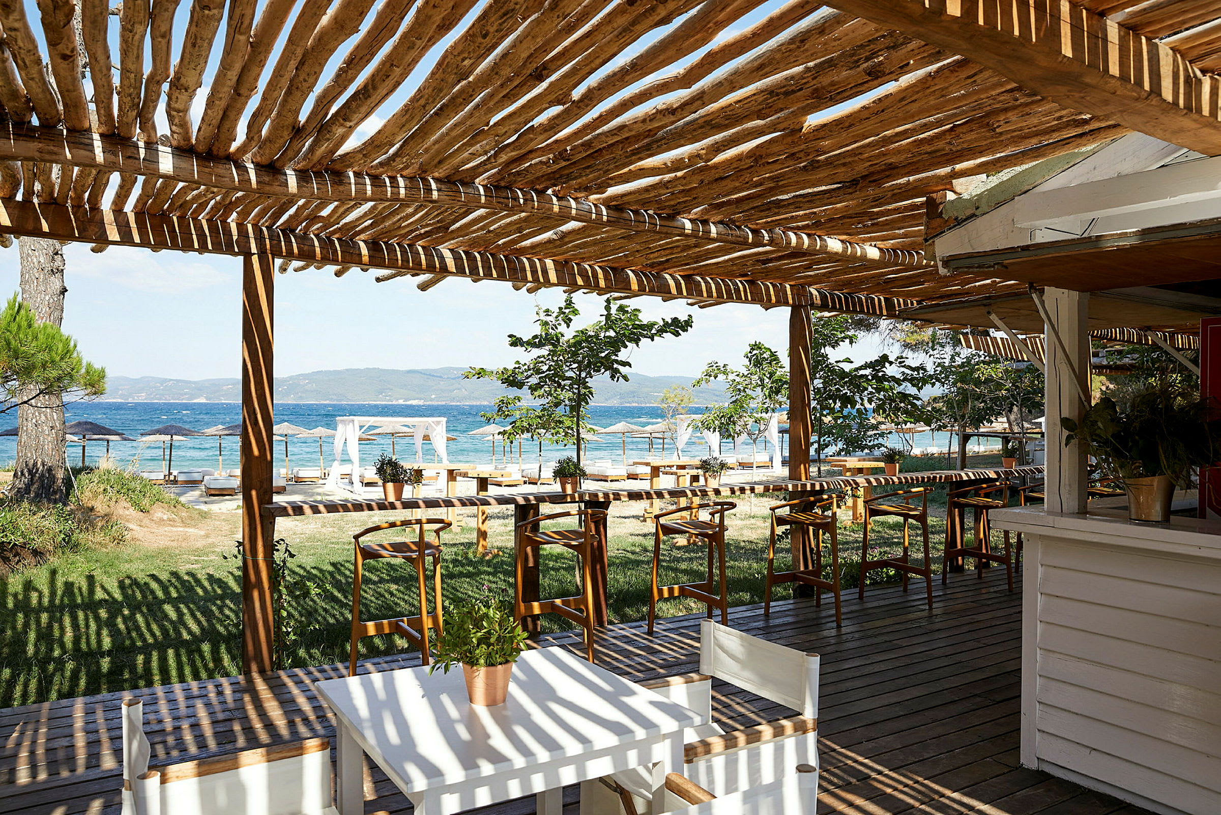En udendørs terrasse med træpergola, borde og stole, med udsigt over havet. (AI-genereret tekst)
