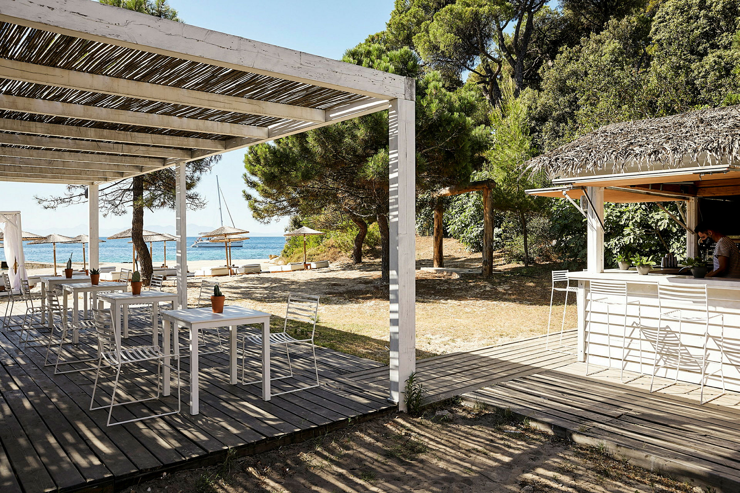 En strandbar med siddepladser under en træpergola, nærliggende parasoller og en skov i baggrunden. (AI-genereret tekst)