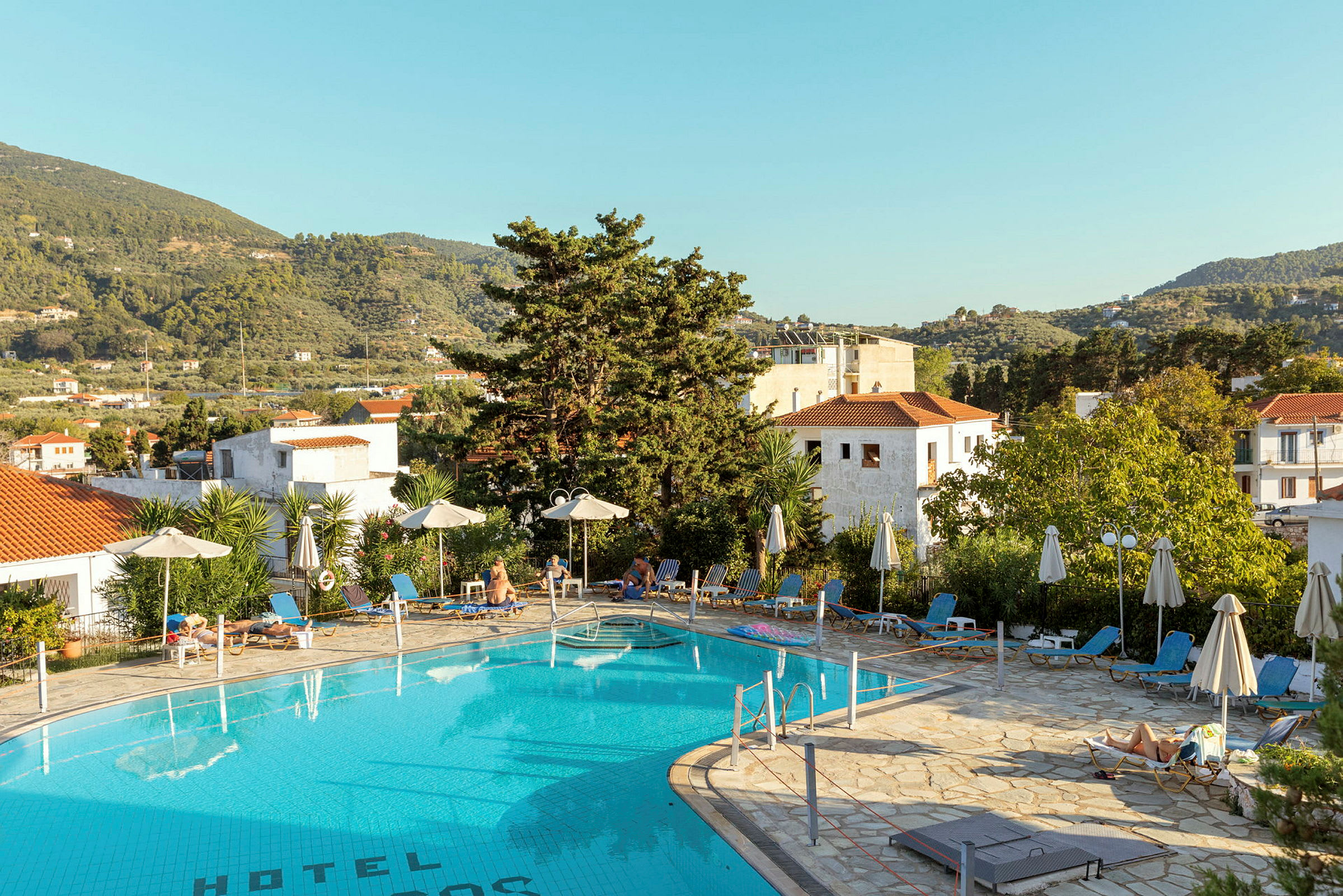 Udendørs pool ved hotel med liggestole og parasoller, omgivet af grønt og bjerge. (AI-genereret tekst)