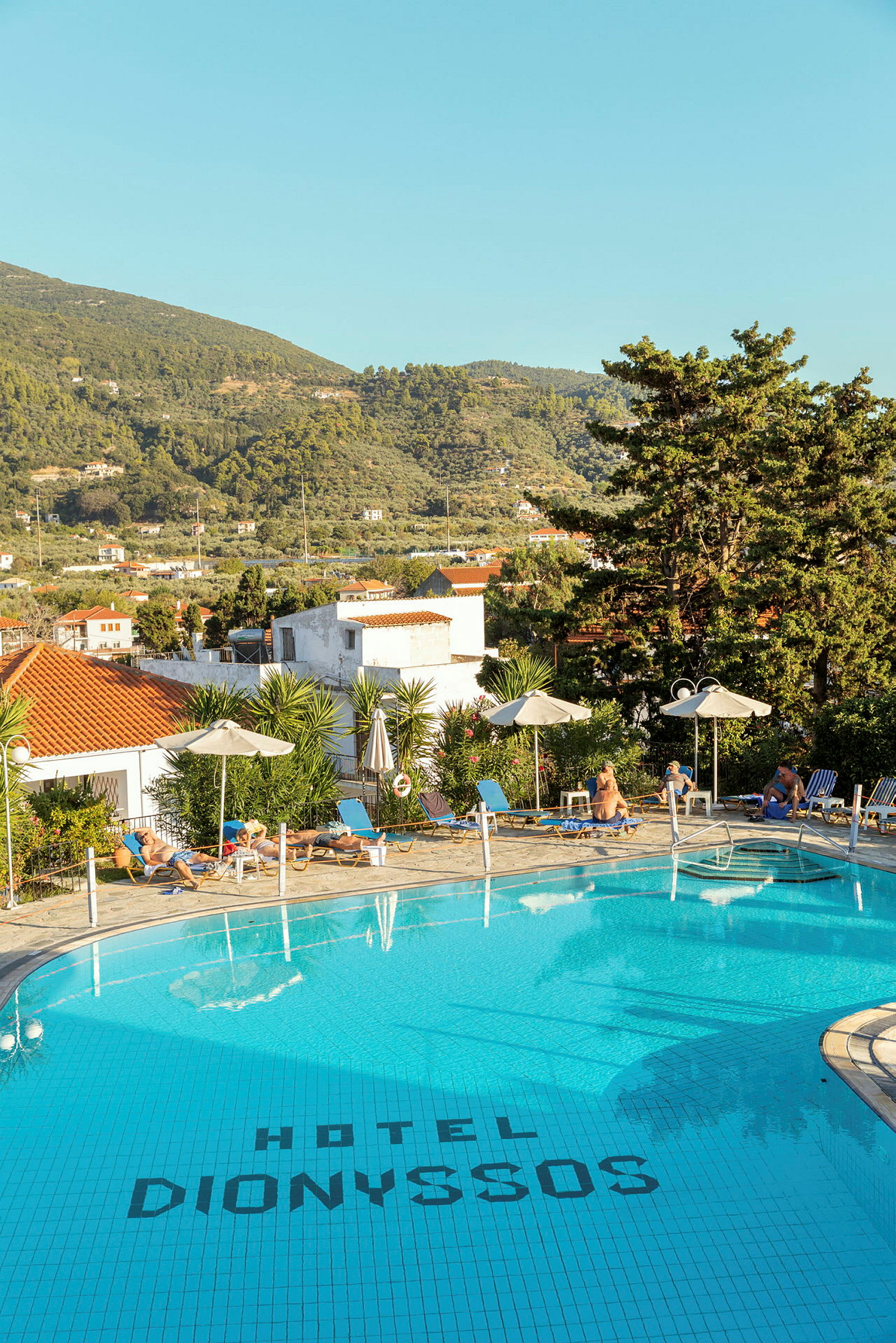 Billede af en pool ved Hotel Dionyssos med liggestole og parasoller rundt om. I baggrunden ses træer og et bjerglandskab. (AI-genereret tekst)