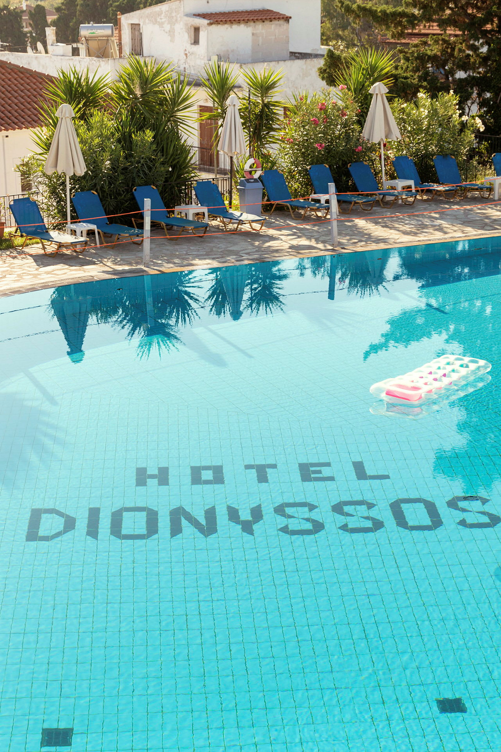 Udendørs pool ved Hotel Dionysos med liggestole og parasoller omgivet af grønne områder. (AI-genereret tekst)
