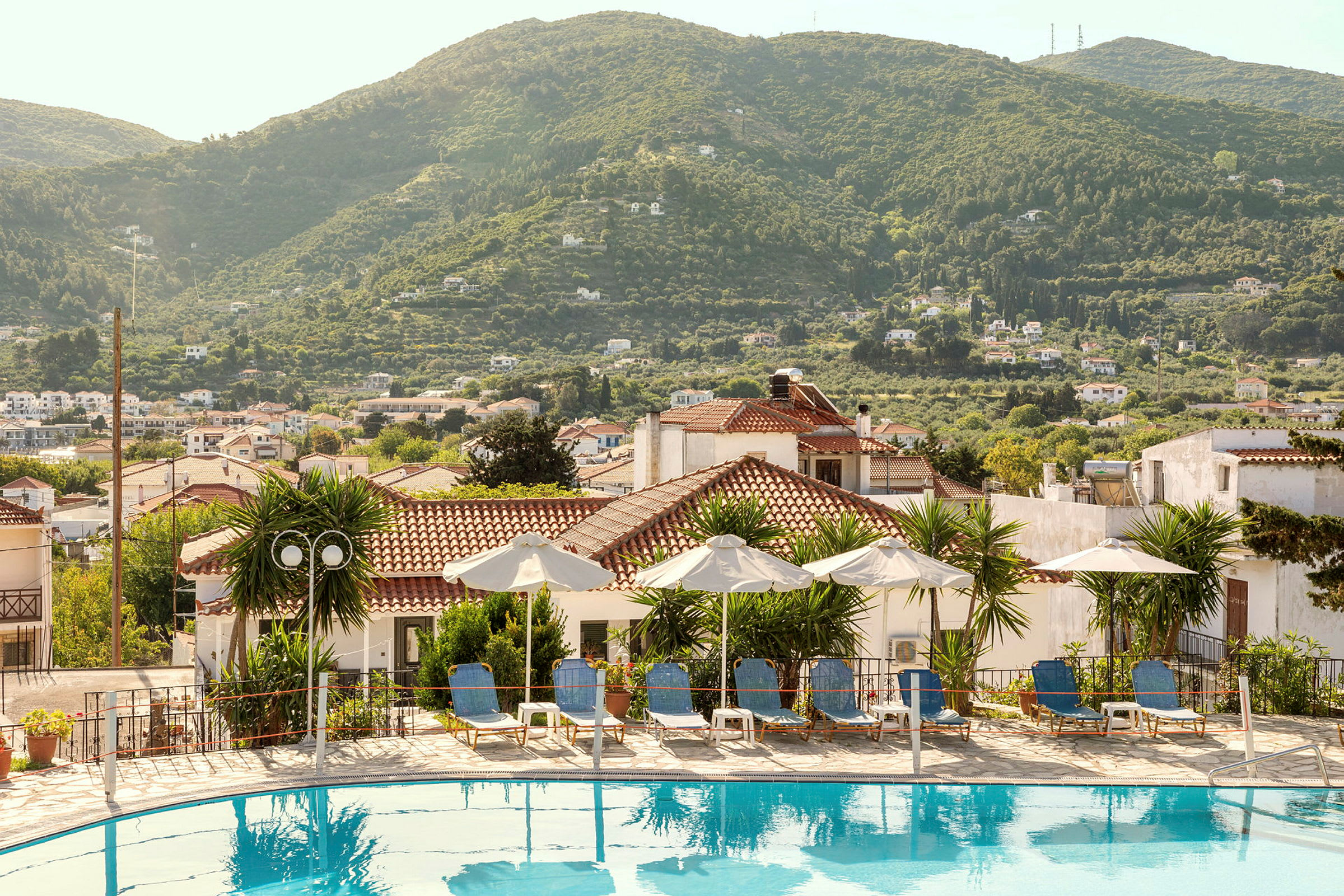 Pool med liggestole og parasoller, omgivet af huse og grønne bakker. (AI-genereret tekst)