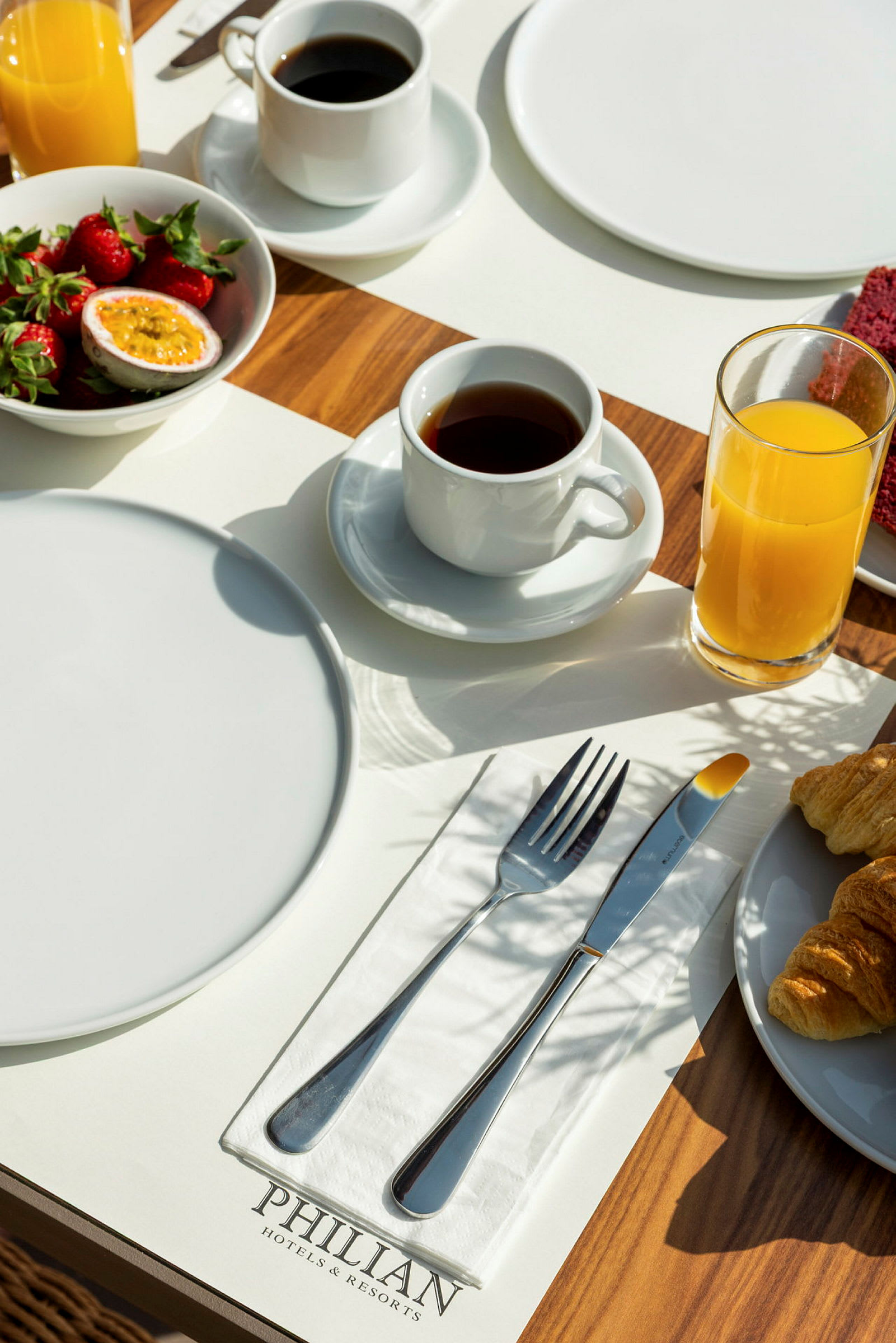 Morgenmadsbord med tallerkener, bestik, kaffe, juice, jordbær og croissanter på en træoverflade. (AI-genereret tekst)