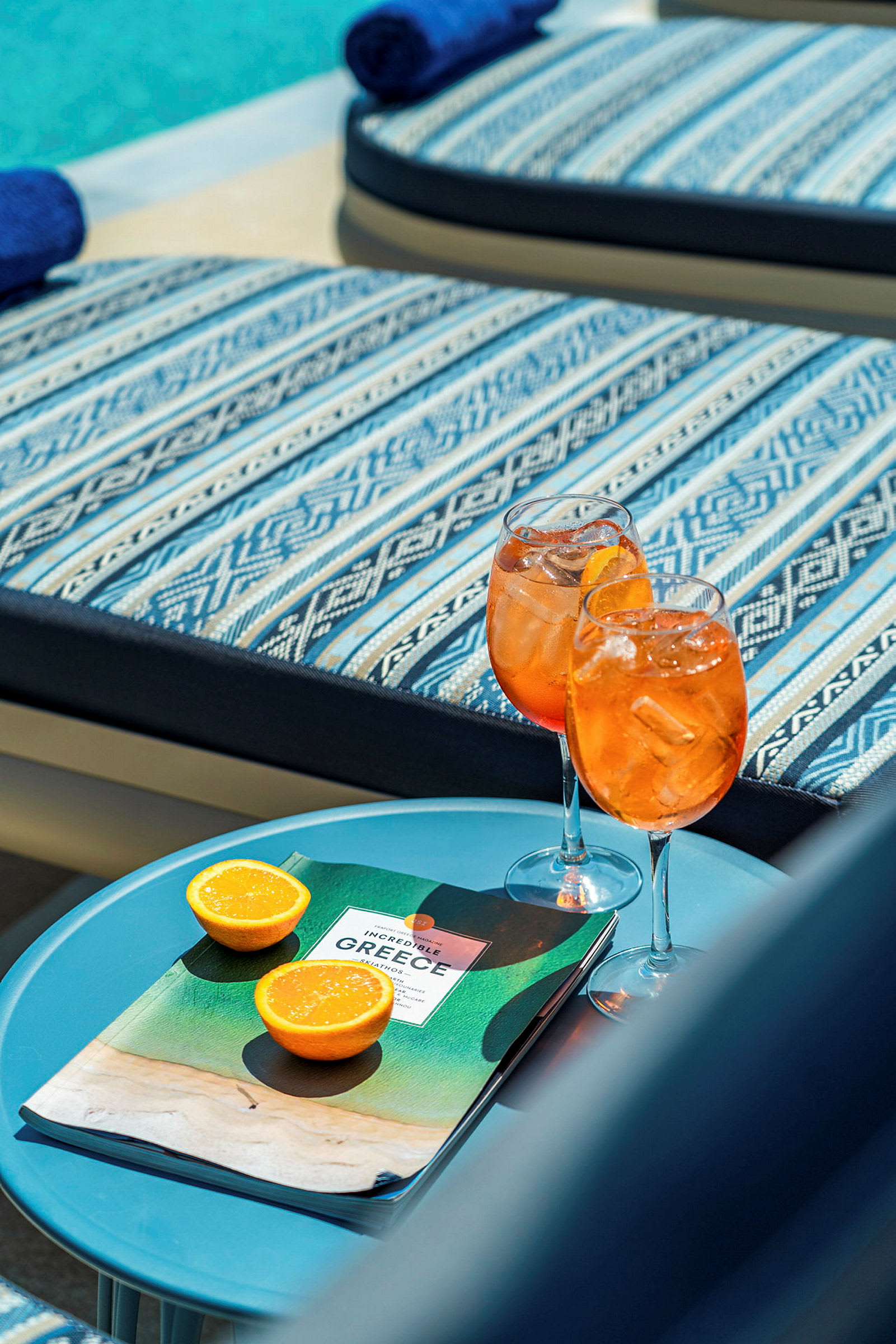 To glas med orange drik, en brochure og to halve appelsiner på et bord ved siden af liggestole. (AI-genereret tekst)