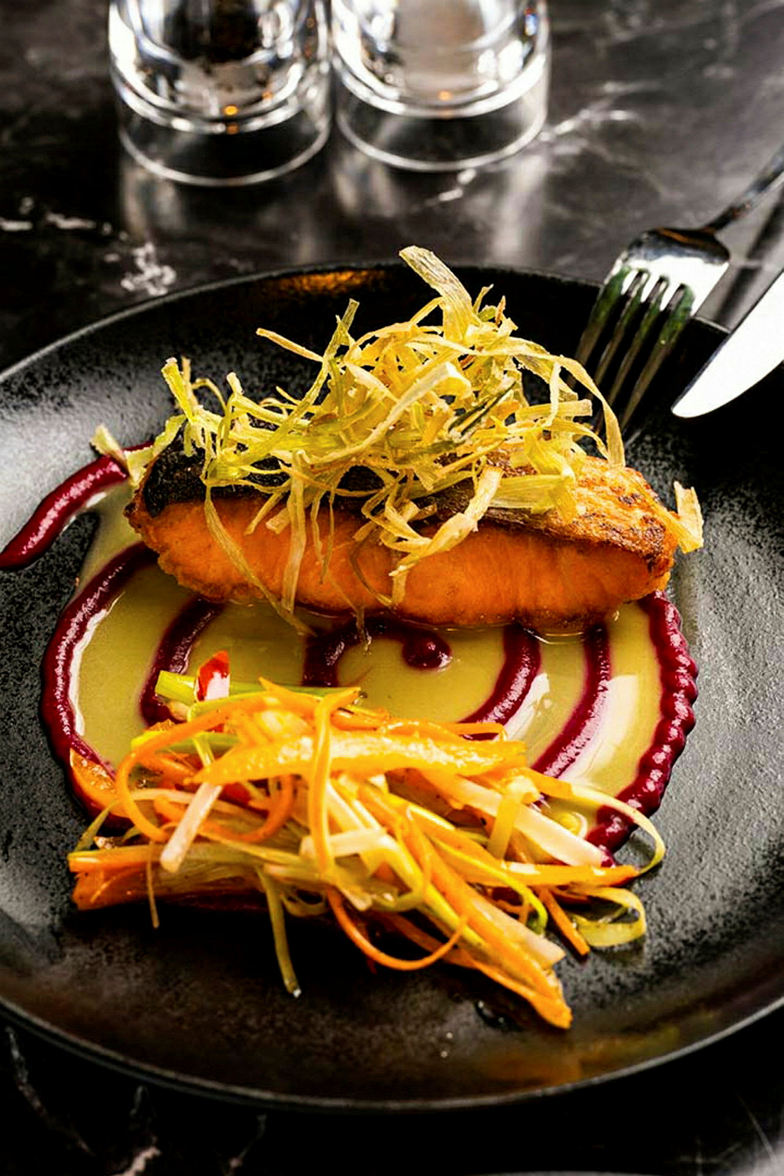 En tallerken med en ret bestående af tilberedt fisk, topping af grøntsager og sauce. Bestik og glas ses i baggrunden. (AI-genereret tekst)