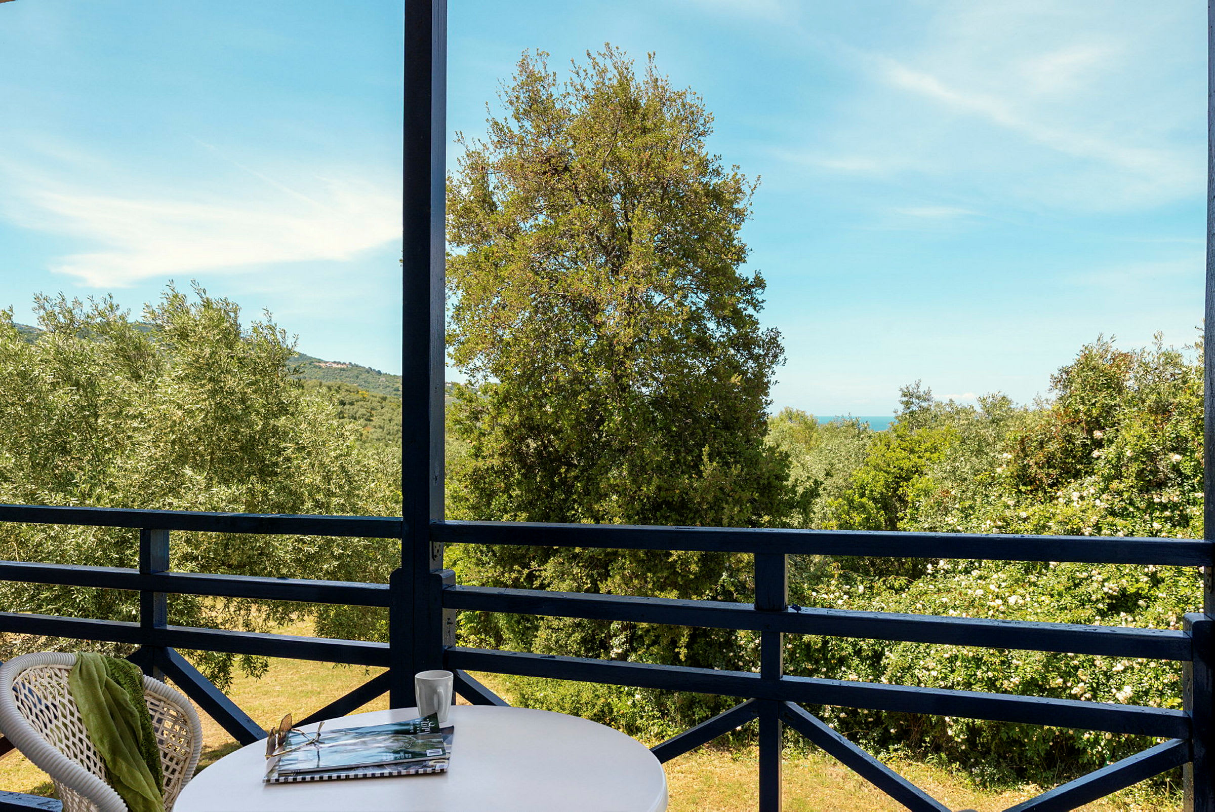 Udsigt fra en balkon med bord og stole mod træer og hav i baggrunden. (AI-genereret tekst)
