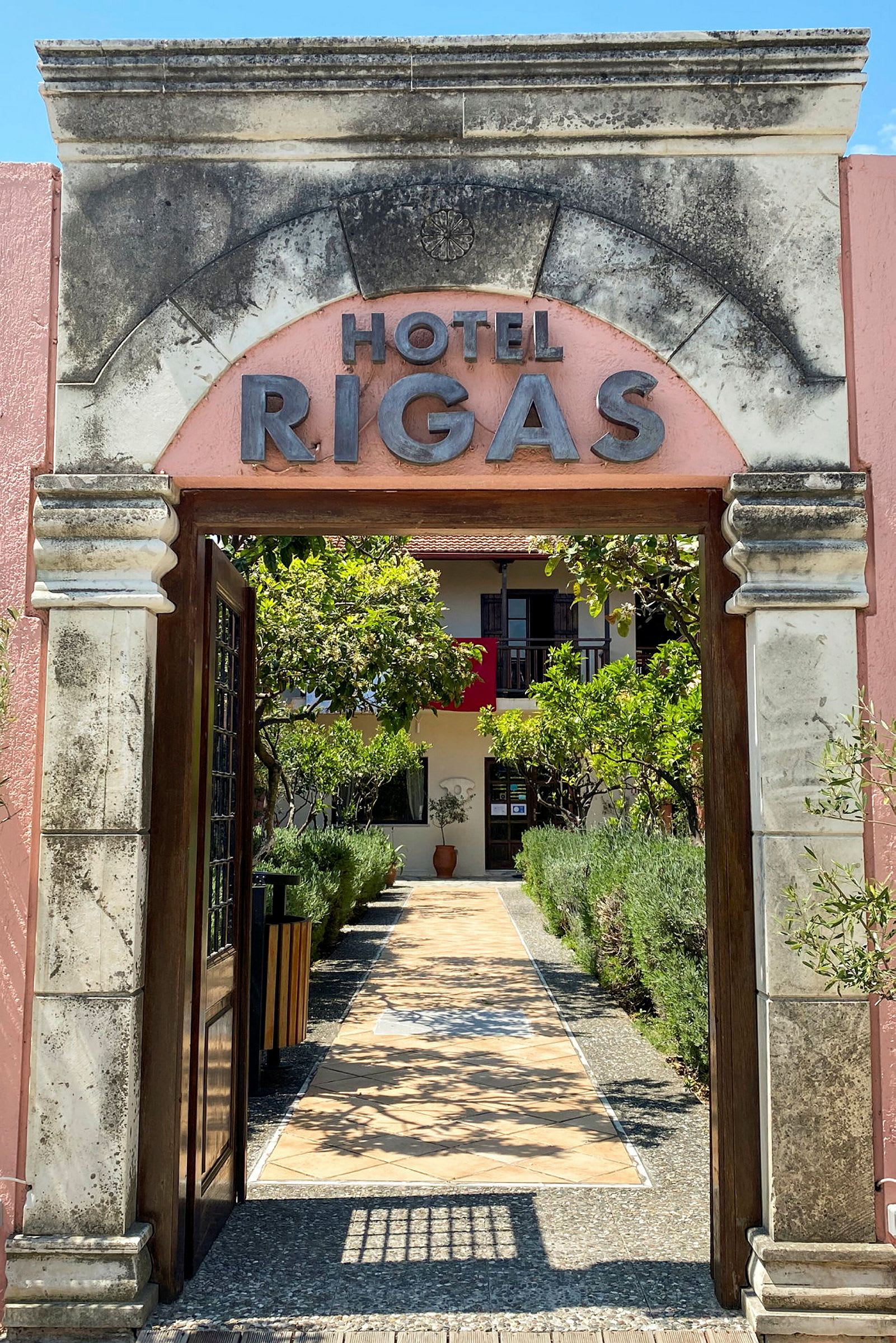 Billede af en hotelindgang med teksten 'Hotel Rigas', omgivet af en have og en sti til døren. (AI-genereret tekst)
