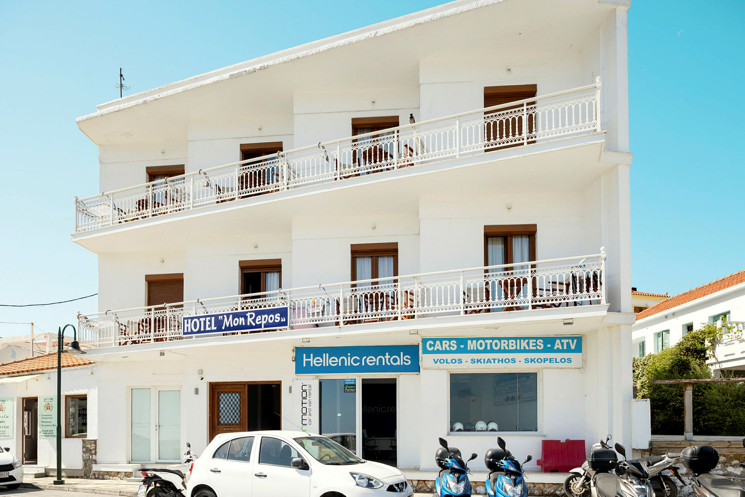 Hvid bygning med skilte for hotel og udlejning af køretøjer. Scootere og en bil er parkeret foran. (AI-genereret tekst)