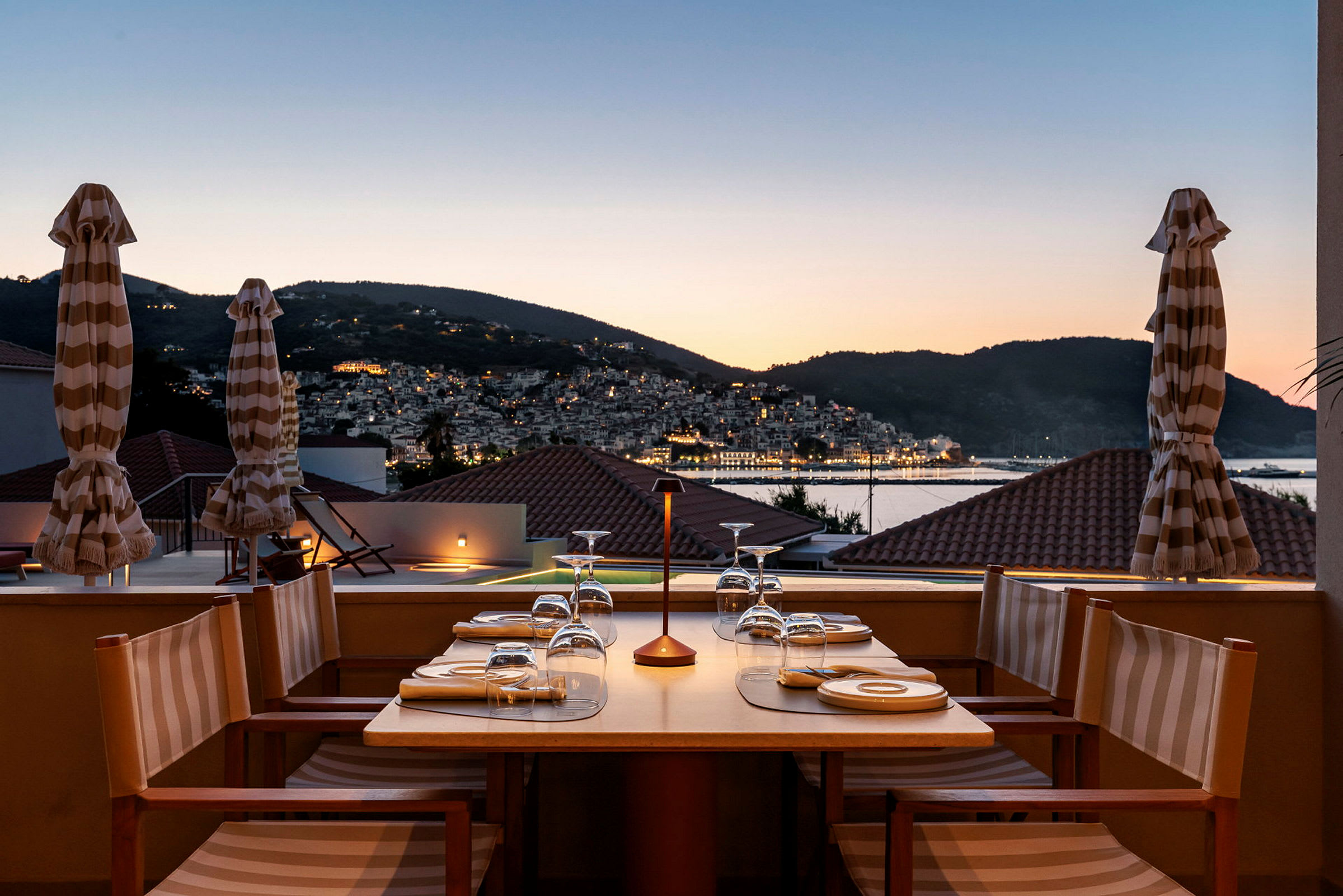 Bord dækket til middag på en terrasse med udsigt over en by og bjerge i skumring. (AI-genereret tekst)
