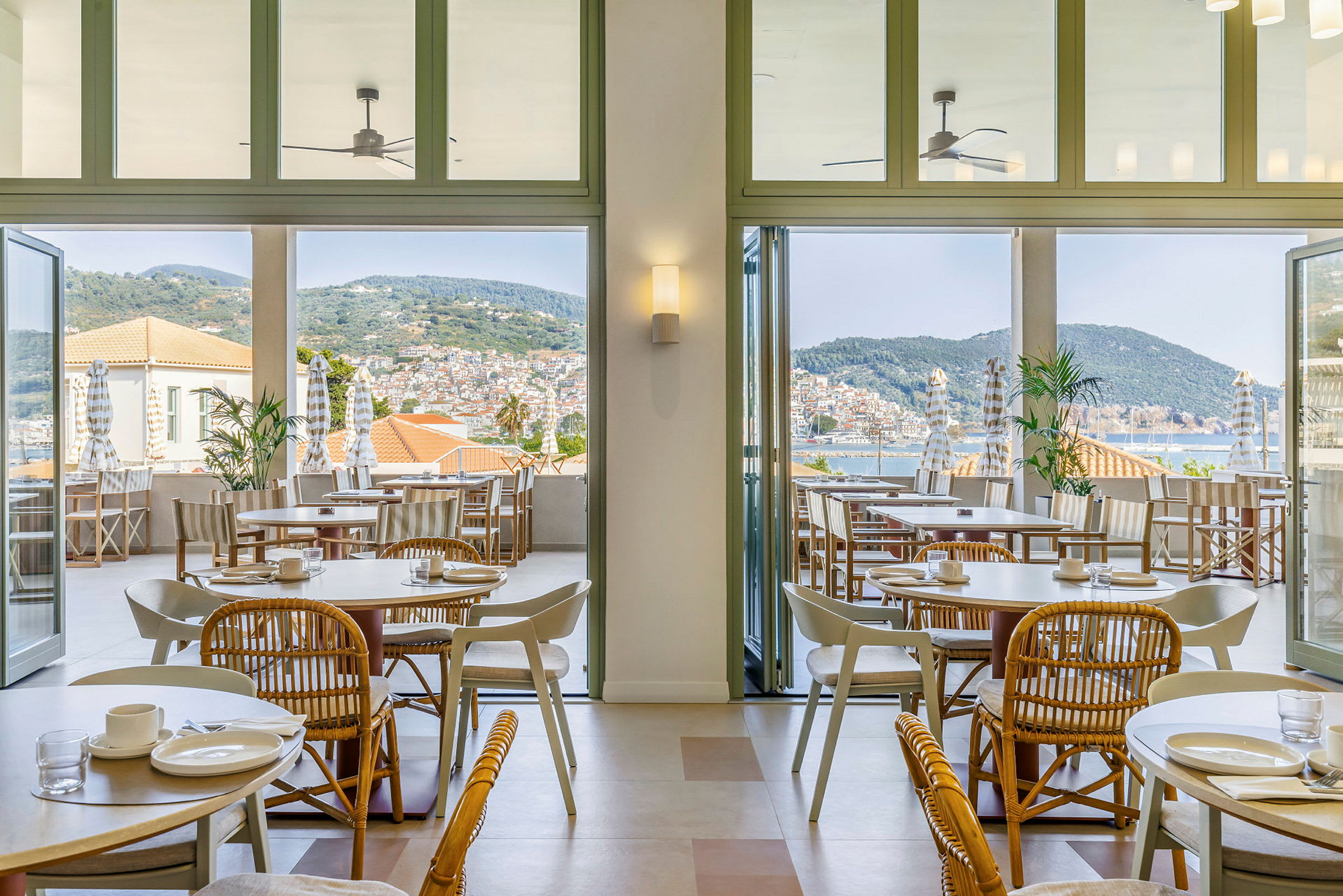 Billede af et lyst og luftigt restaurantområde med borde og stole indendørs samt en terrasse med udsigt over havet og byen. (AI-genereret tekst)