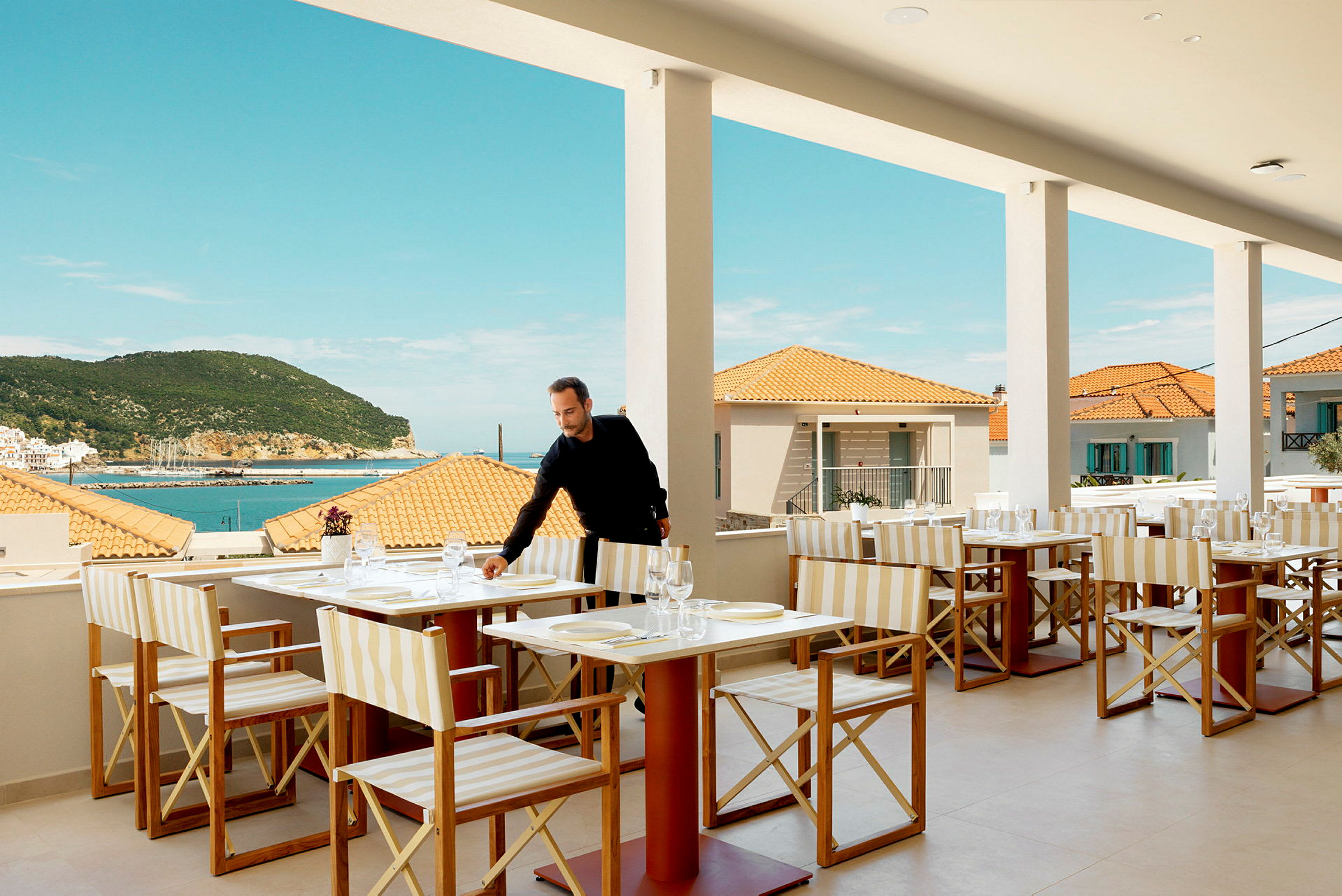 Restaurant med bordopstilling, udsigt over havet og terrasse, med baggrund af terrakottatag og en frodig bakke. (AI-genereret tekst)